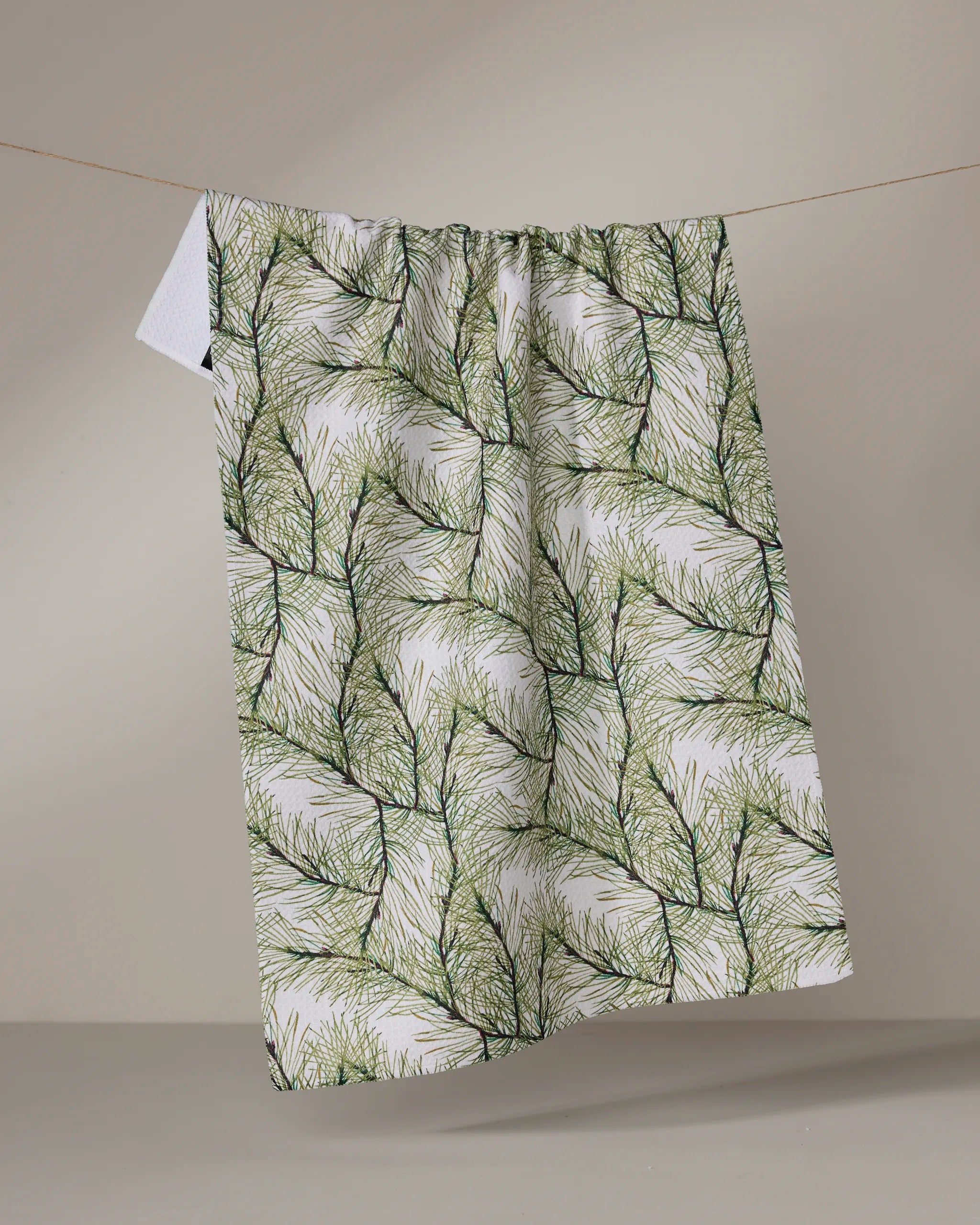 The Geometry Pinewell recycled kitchen towel, featuring a green pine needle pattern, hangs naturally on a line against a beige background, highlighting its bold botanical design.