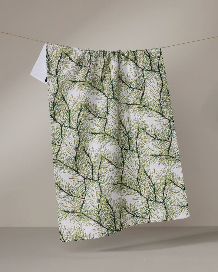 The Geometry Pinewell recycled kitchen towel, featuring a green pine needle pattern, hangs naturally on a line against a beige background, highlighting its bold botanical design.