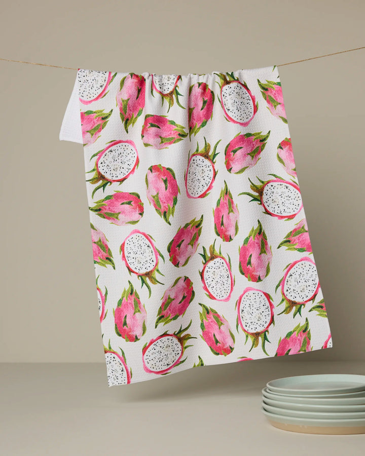 The Geometry Pink Dragon tea towel, featuring a vibrant dragon fruit design, hangs above a stack of white plates on a beige background, bringing both charm and function to your kitchen decor.