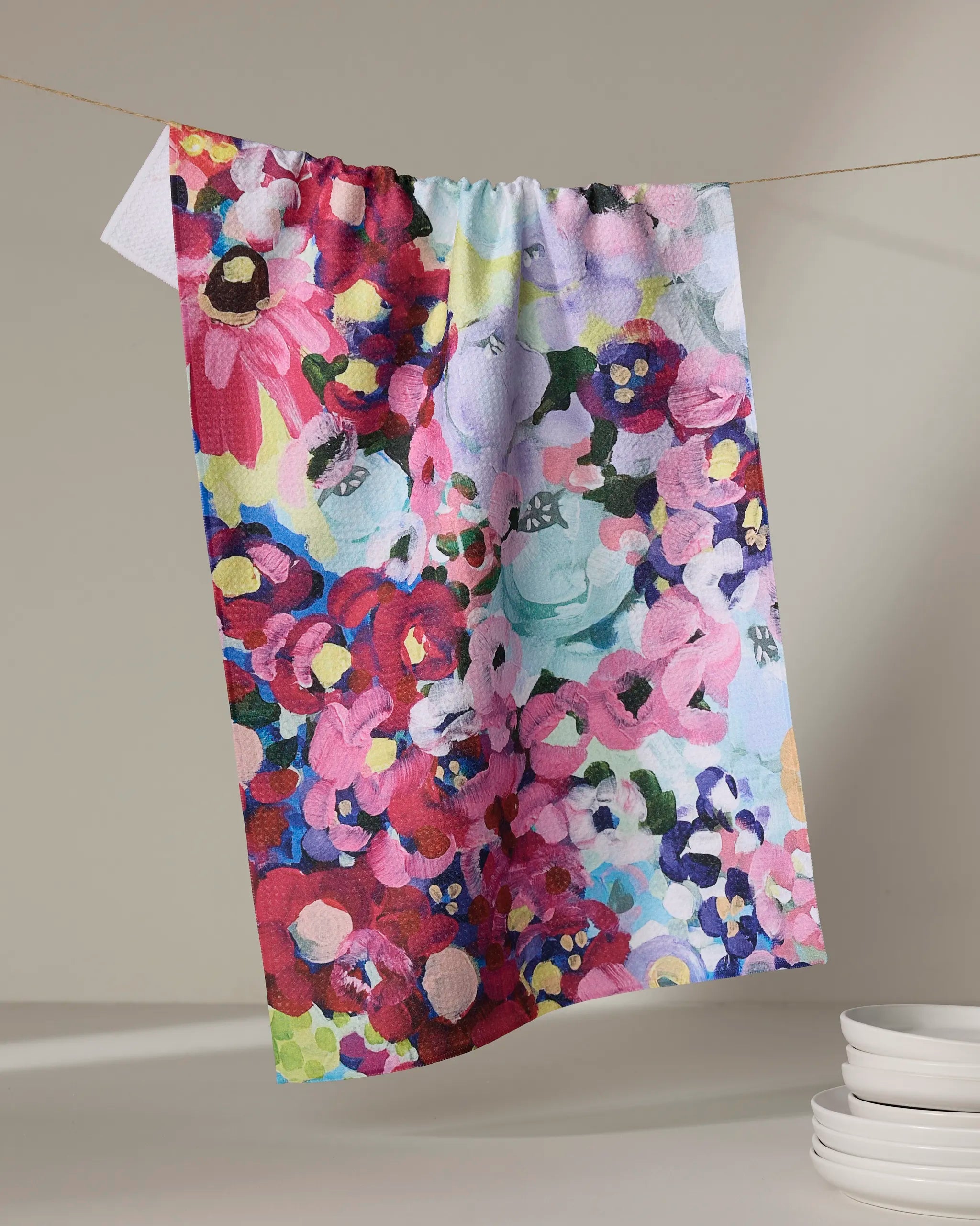 The Pink Fields of Joy dish towel by Ania Zwara Art, featuring a vibrant floral pattern, hangs on a line against a neutral background with a stack of white dishes positioned below and to the right.