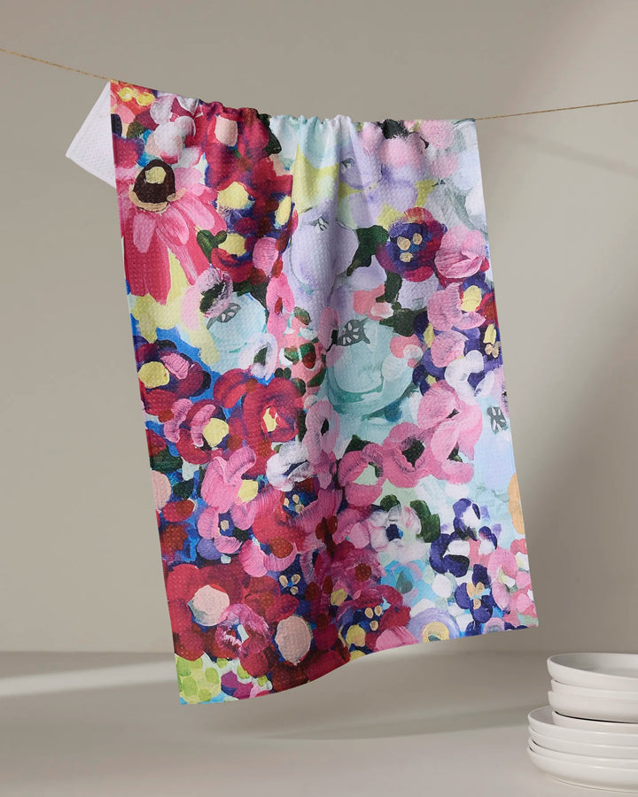 The Pink Fields of Joy dish towel by Ania Zwara Art, featuring a vibrant floral pattern, hangs on a line against a neutral background with a stack of white dishes positioned below and to the right.