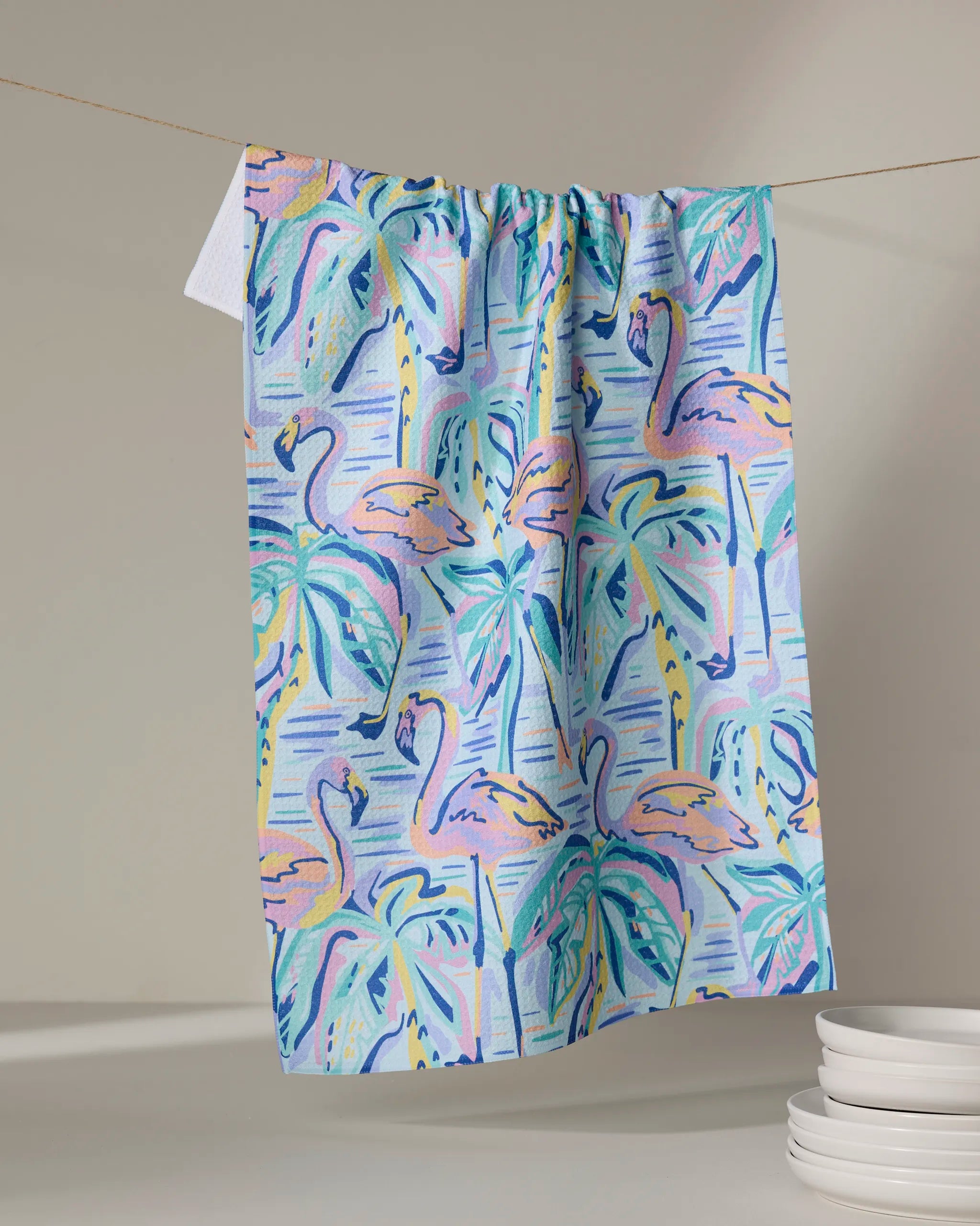 The Pink Flamingo tea towel by Masha Volnova, featuring a pastel pink, blue, and green flamingo and palm tree design, hangs on a line beside stacked white plates against a soft neutral background.