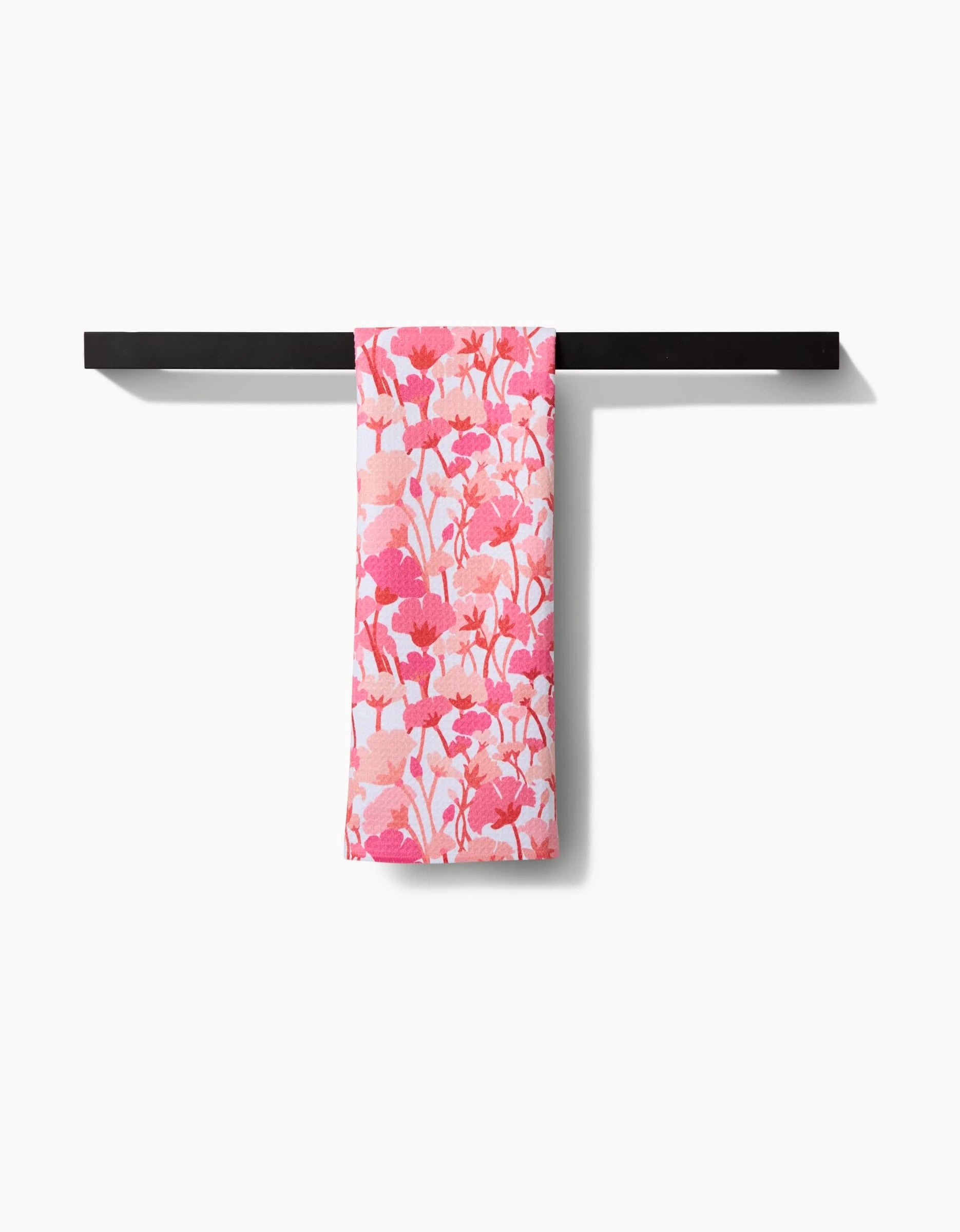 The Pink Florals tea towel by Katie OShea, featuring a pink and white floral design on Geoweave fabric, is neatly draped over a black towel bar against a plain white background.
