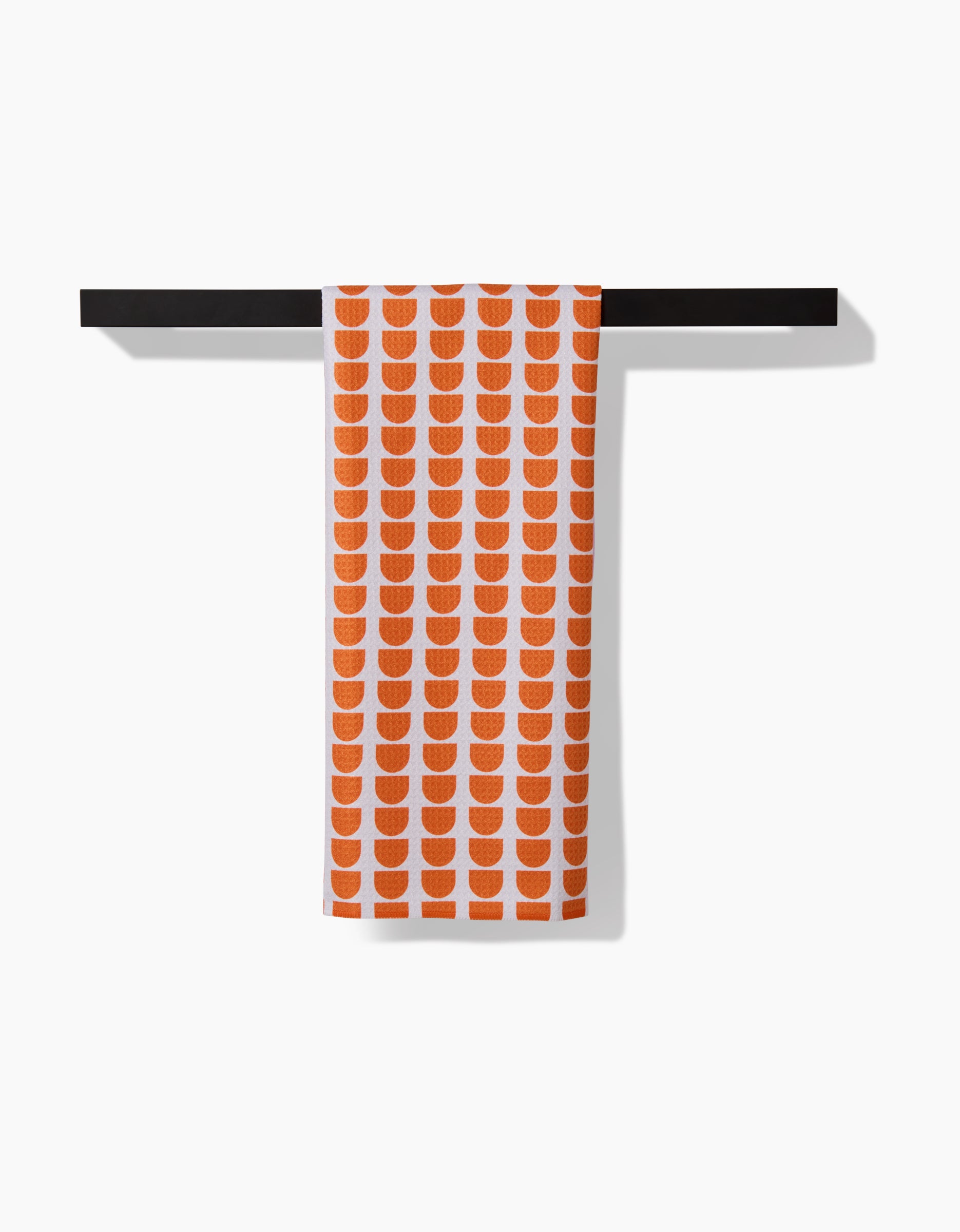 A white towel named Pink Illusion by Happy Hemera, crafted from ultra-absorbent Geoweave™ fabric with a repeating pattern of orange semicircles, hangs neatly over a black rod against a white background.