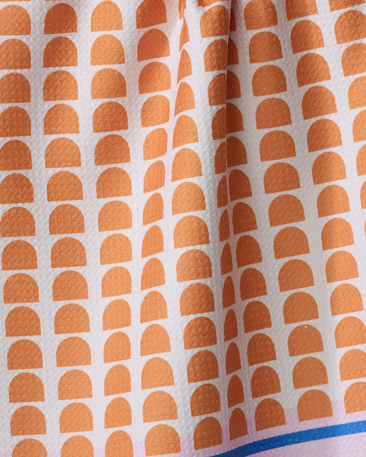 A detailed view of Happy Hemera's Pink Illusion tea towel shows textured fabric with orange semicircles on white, a blue stripe at the bottom, and ultra-absorbent quality—perfect for any kitchen.