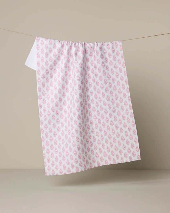 The Geometry Pink Poppy tea towel, made from ultra-absorbent recycled materials, features a repeating pink floral teardrop pattern and hangs on a thin string against a plain beige background.
