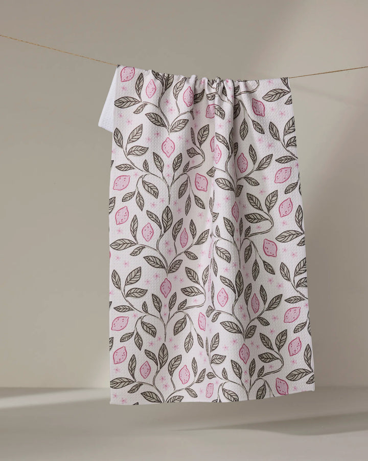 The Pink Summer Lemons tea towel by Teri the Butter, made from recycled materials, features pink and green leaves and hangs from a clothesline against a neutral background.