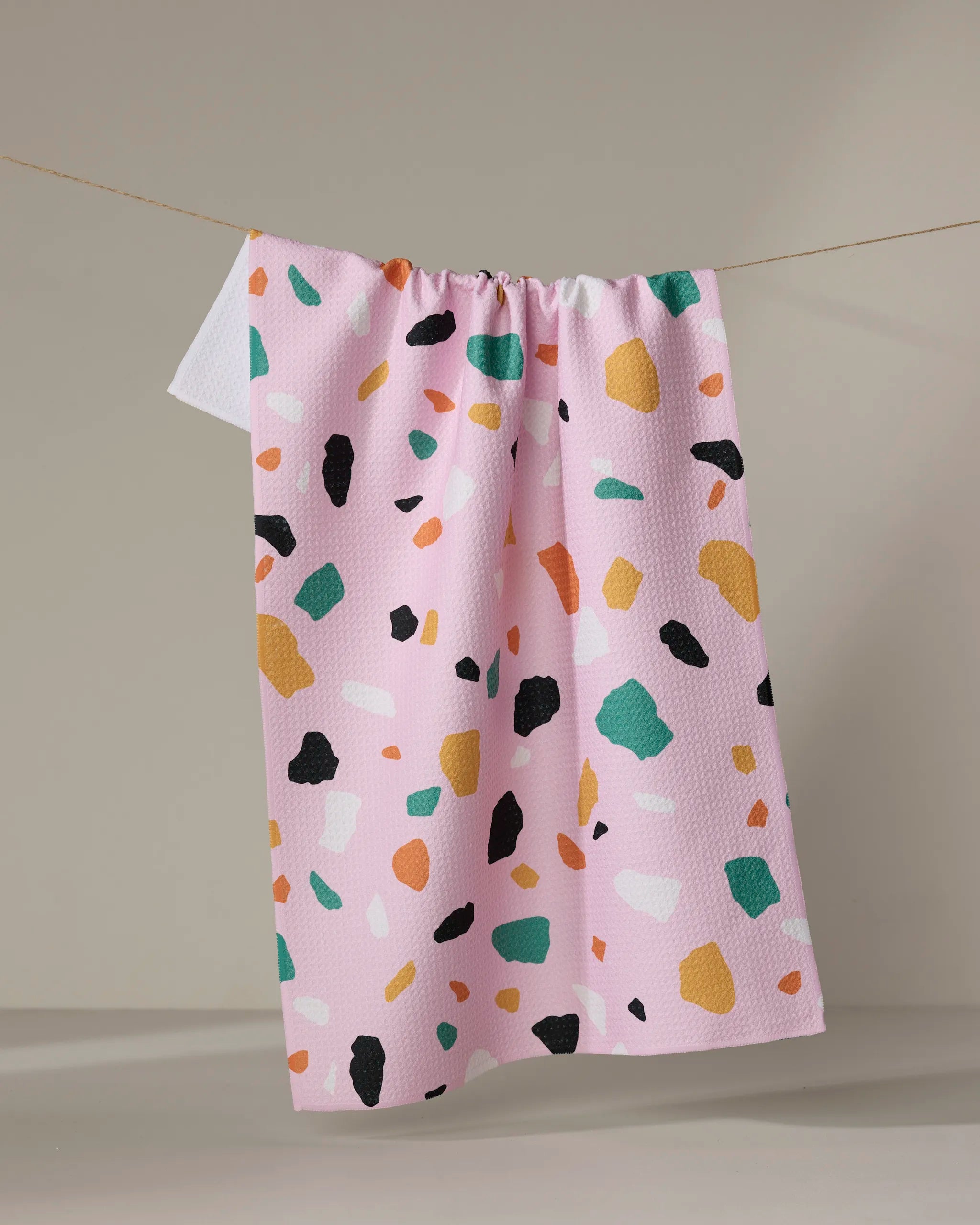 The Megan Roy Pink Terrazzo kitchen towel features a pink textured fabric with green, orange, yellow, white, and black terrazzo shapes. This eco-friendly accent brightens any kitchen with its playful, modern design.