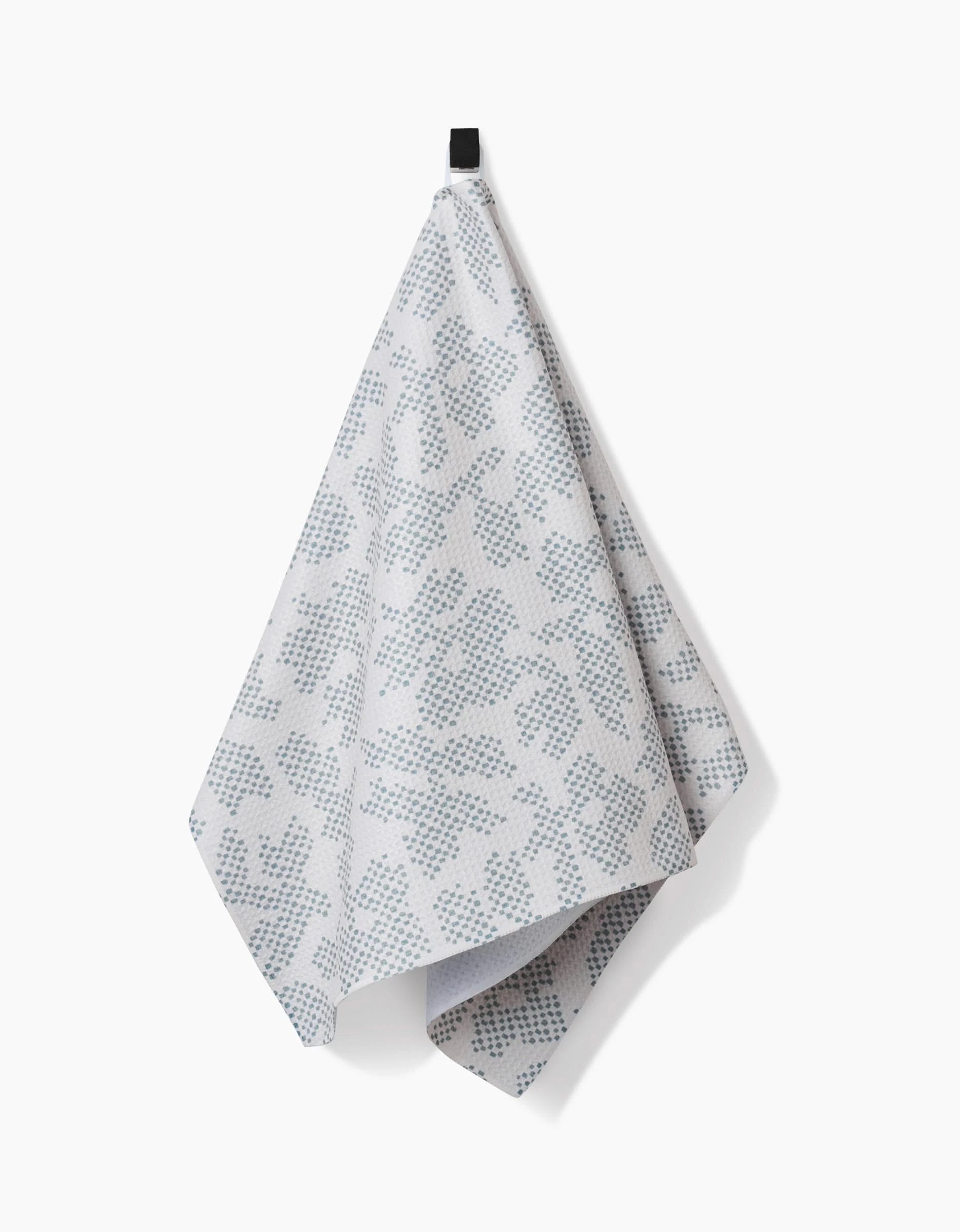 The Pixie Sage tea towel by Studio Jeannot, in light gray with a blue geometric pattern and made from Geoweave™ fabric, hangs from a black hook against a plain white background.