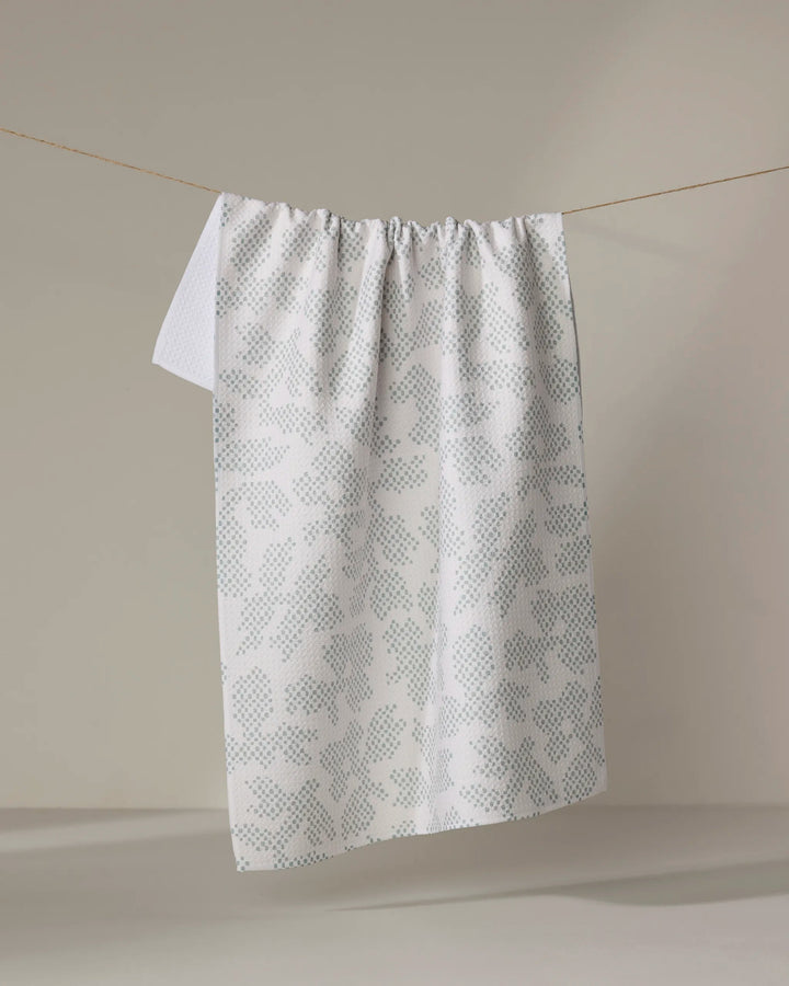 The Pixie Sage tea towel by Studio Jeannot, featuring a light blue-grey dotted pattern on white and crafted from recycled materials, hangs on a clothesline against a neutral beige background.