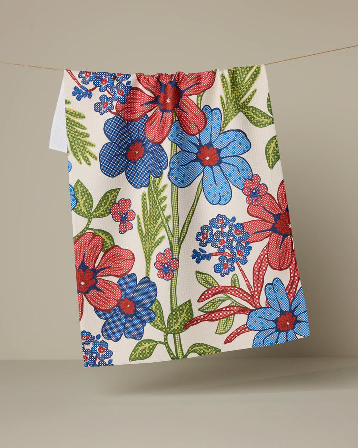 The Plaid Floral tea towel by Geometry, featuring bold red, blue, and green florals and made from recycled materials, hangs on a line against a plain beige background.