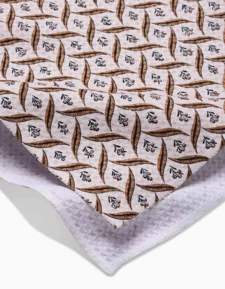 A close-up of Hufton Studios Plated Fall Berry Natural fabric, featuring a repeating leaf and floral design in brown, blue, and black on white, made from recycled materials and layered over textured white cloth.