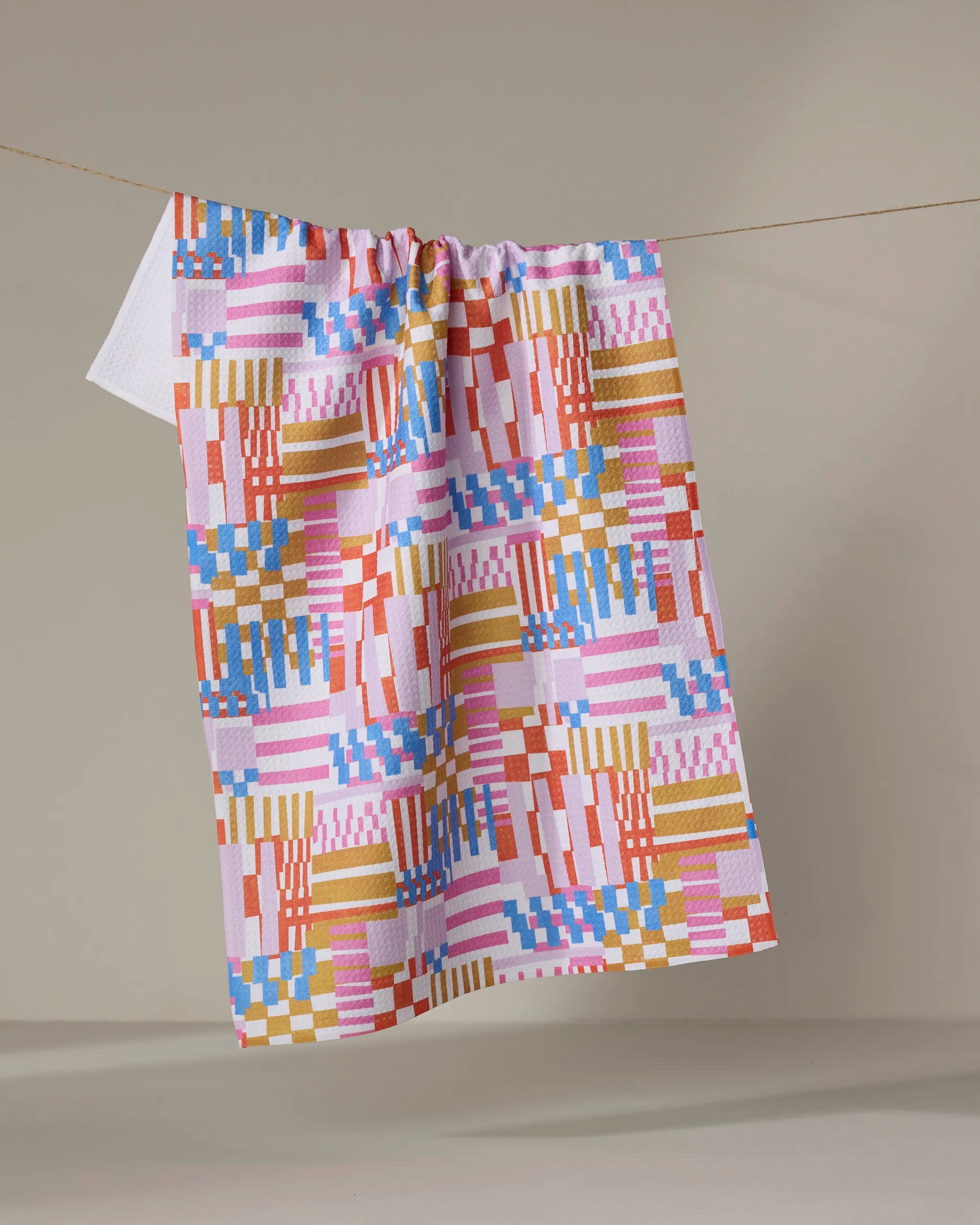 The "Playful Stripes" recycled kitchen towel by Claire Iglesias, featuring red, pink, blue, yellow, and white geometric squares and stripes, hangs on a string against a neutral background.