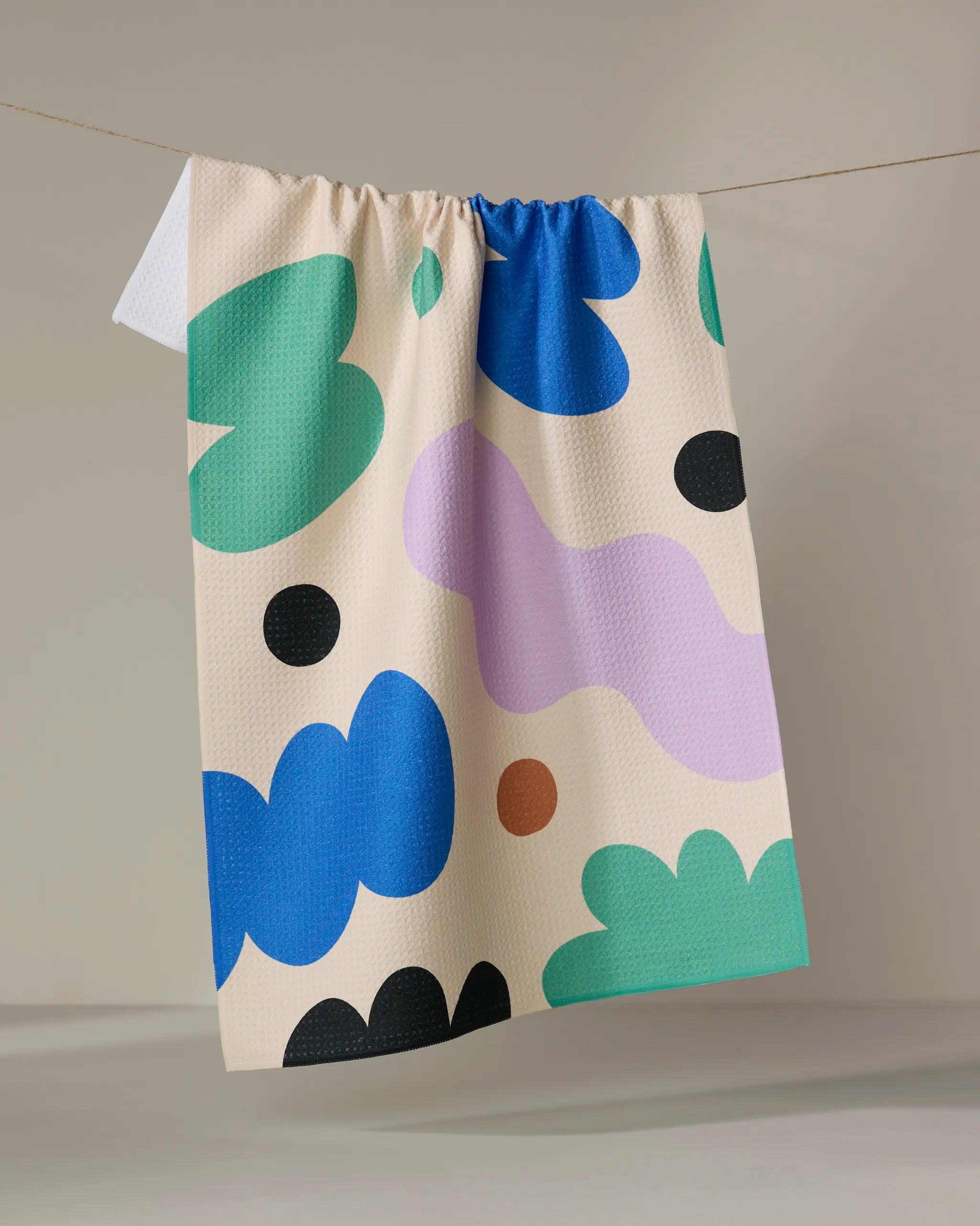 The "Playground" dish towel by Beetle Ink Co. showcases abstract shapes in green, blue, black, purple, and brown, hanging on a clothesline against a plain gray background.