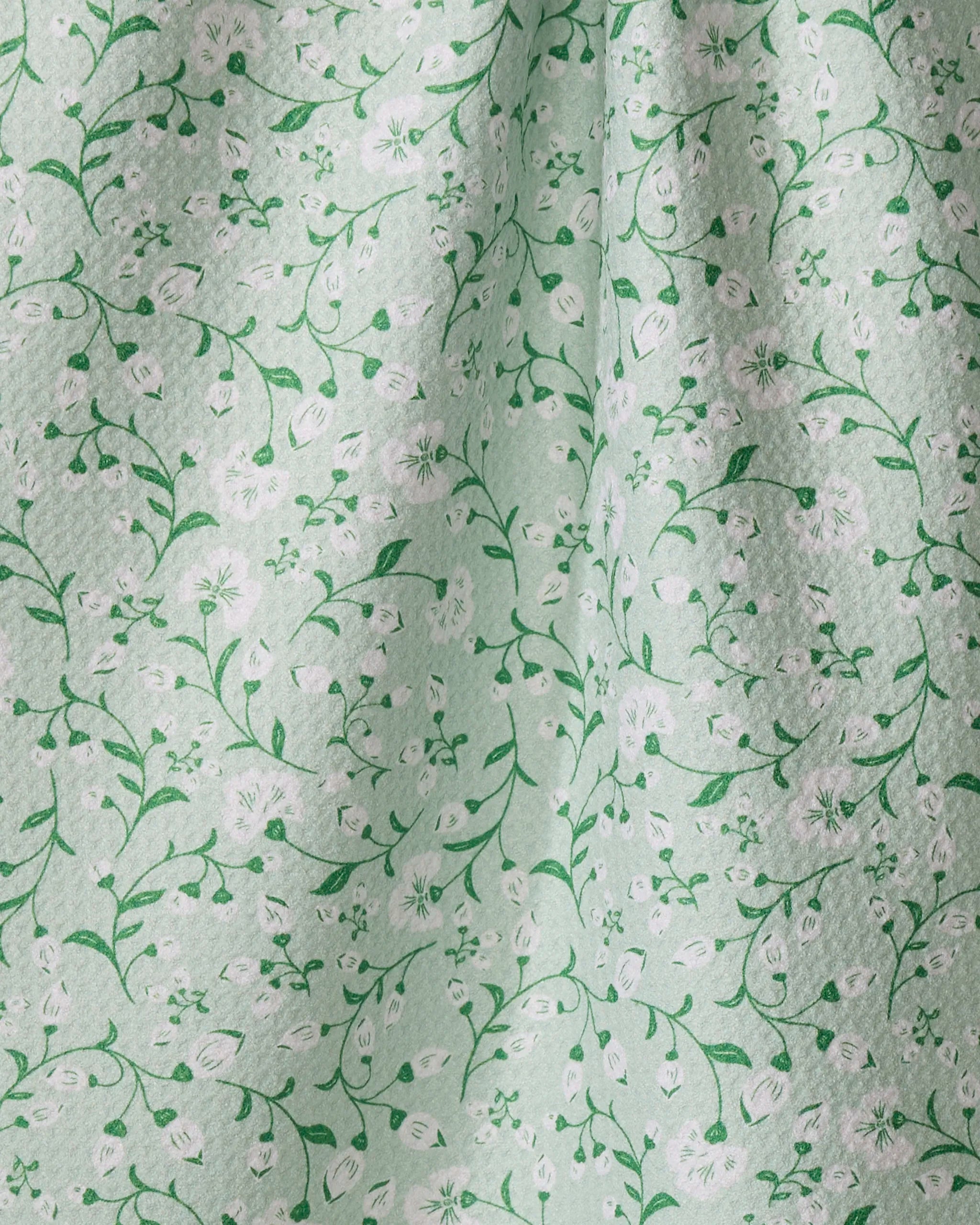 The Plumbago Bloom kitchen towel by Danelys Sidron features a light green, recycled fabric with a subtle texture and an elegant pattern of green vines and white flowers. Its gentle folds create light and shadow on the quick-drying surface.