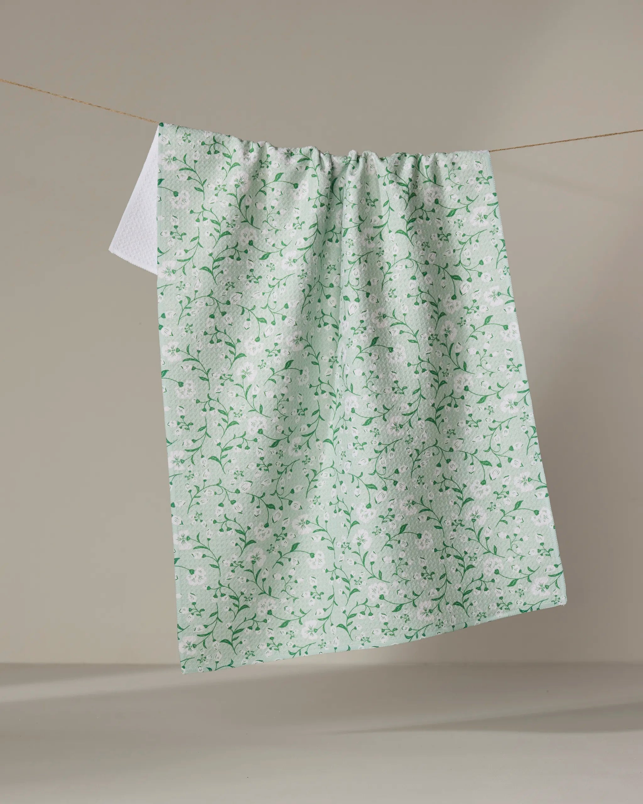 The Plumbago Bloom tea towel by Danelys Sidron, featuring a light green and white floral pattern, hangs on a clothesline against a plain beige background.