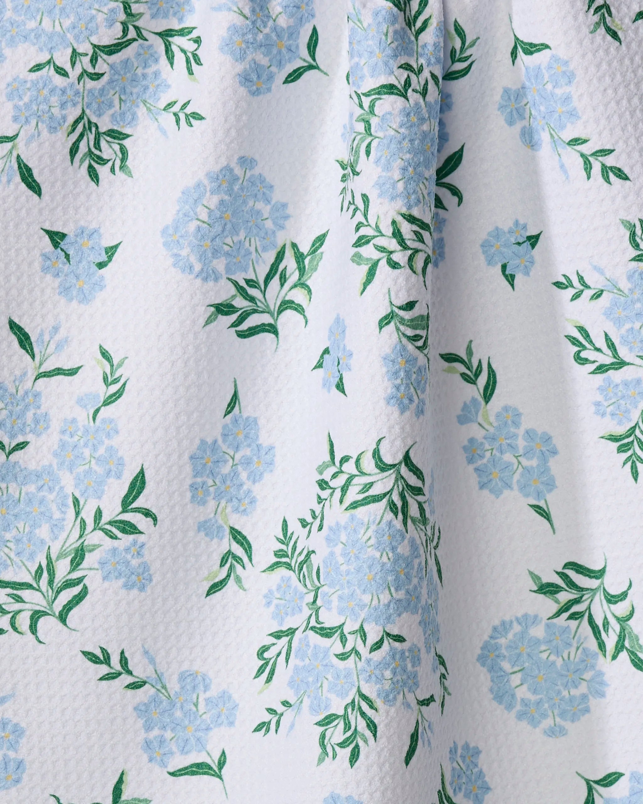 The Plumbago tea towel by Danelys Sidron features white textured fabric with light blue flower clusters and green leaves. Gentle folds enhance the floral design, creating a beautiful and practical addition to your kitchen.