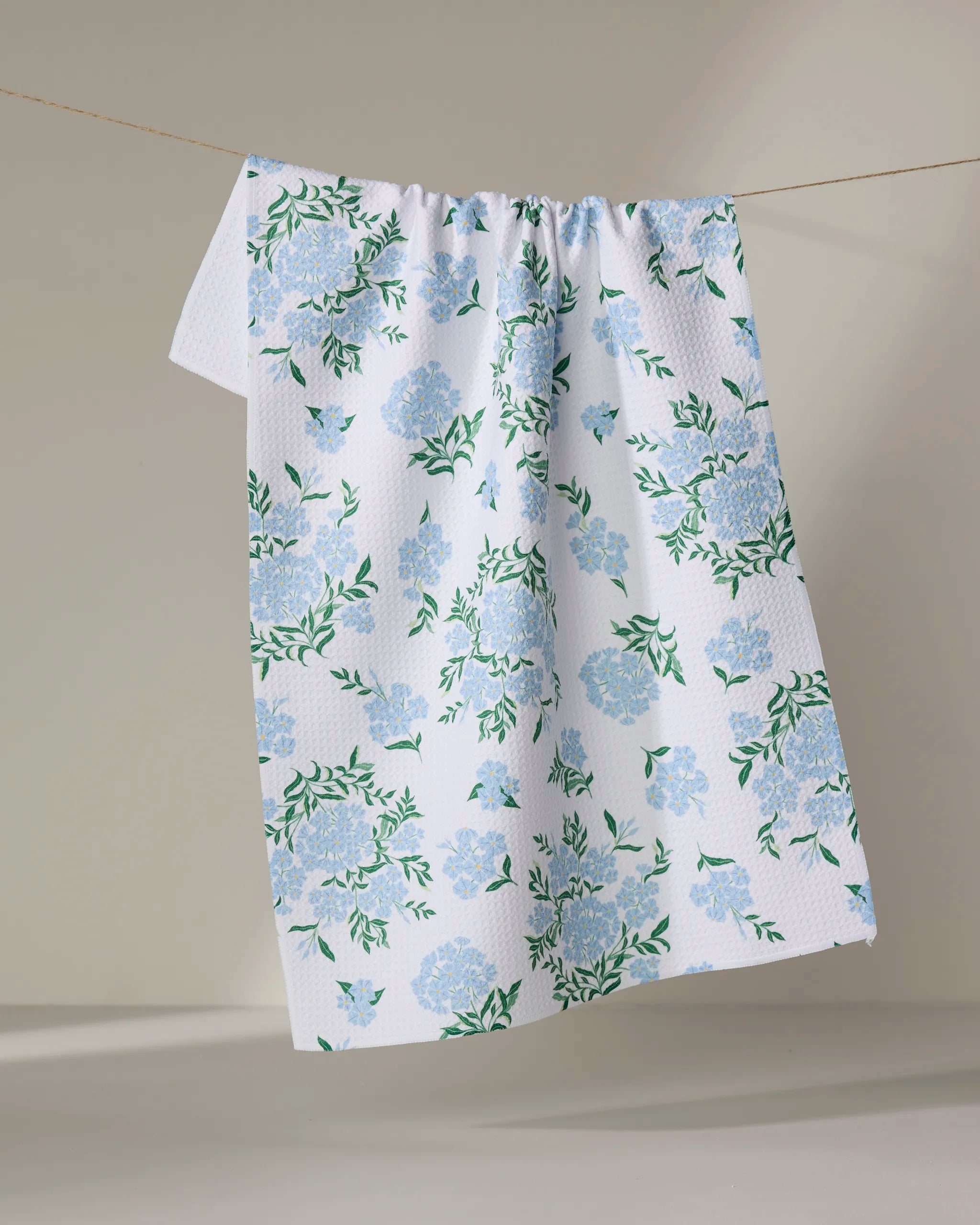 The Danelys Sidron Plumbago tea towel, featuring a blue and green floral design, is draped over a clothesline against a neutral background.