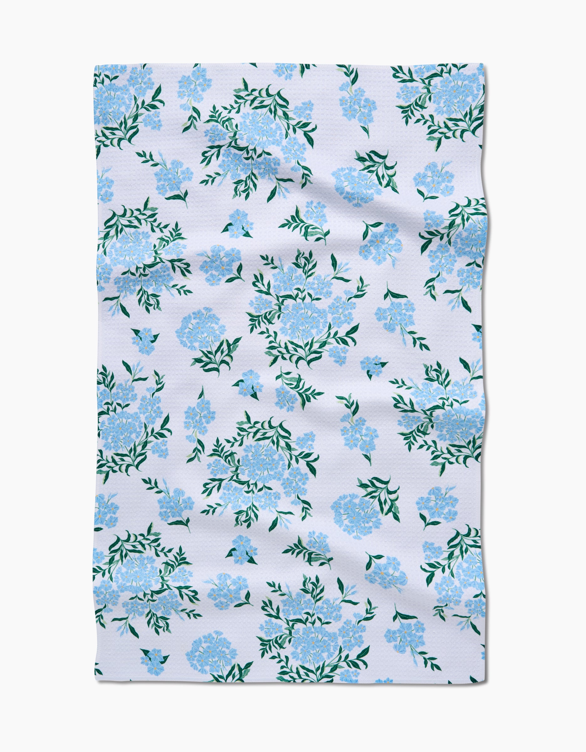 The Plumbago by Danelys Sidron is an ultra-absorbent white fabric made from recycled materials, featuring a repeating blue flower and green leaf pattern. It’s displayed slightly wrinkled and laid flat.
