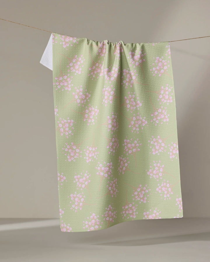 The Pocket of Posies kitchen towel by Sarah Reed, in light green with small pink and white floral patterns, hangs over a clothesline against a neutral background.