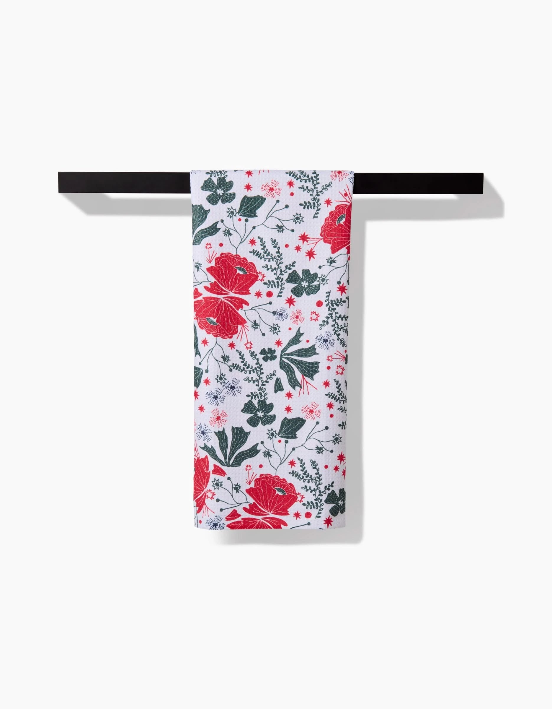 The Ash Cascade Poinsettia Bramble tea towel, made from Geoweave fabric and featuring a red and green floral pattern, is shown draped over a black towel bar against a white background.