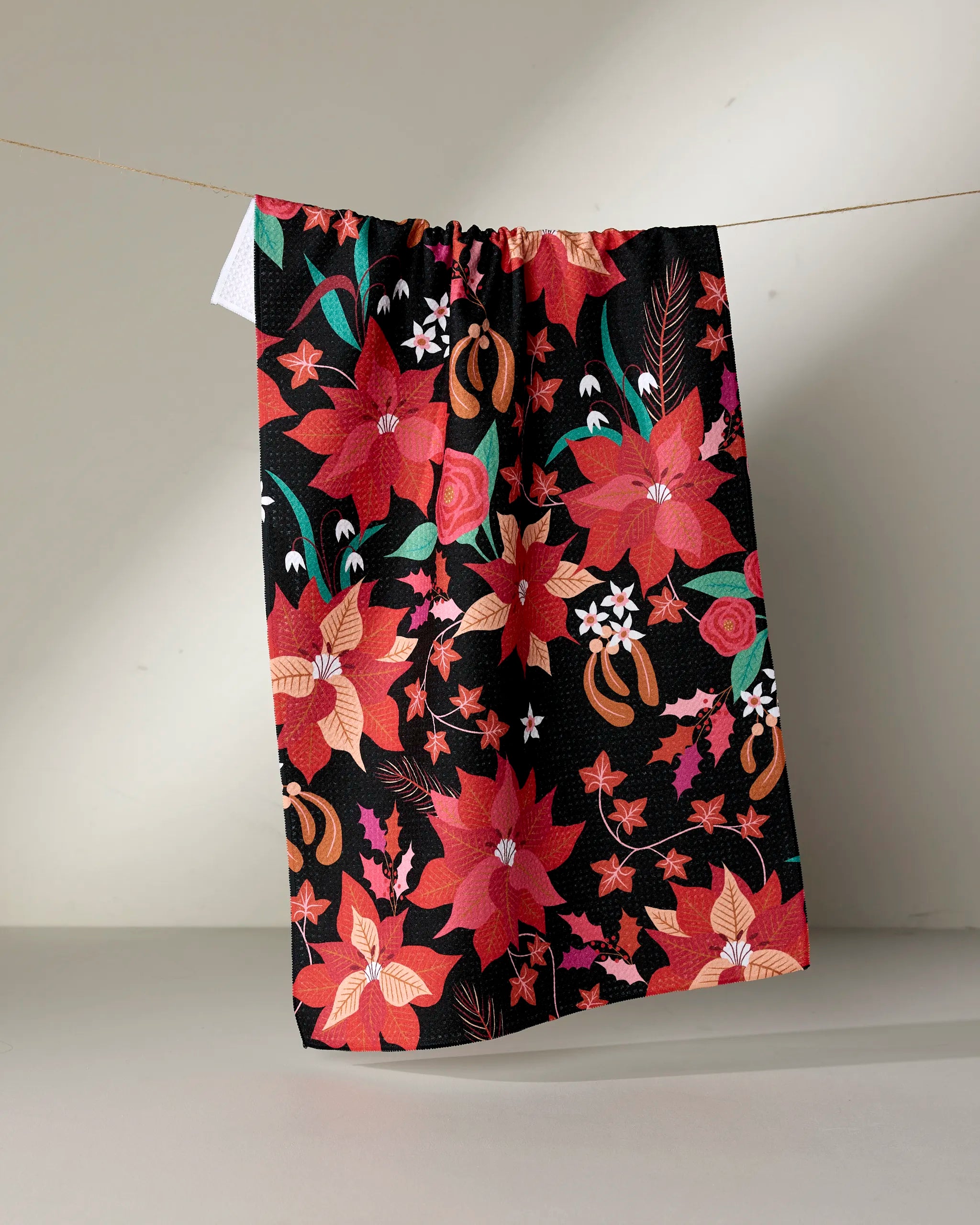 The Rachel Parker "Poinsettia" kitchen towel, featuring a vibrant floral design with red, pink, and orange flowers plus green leaves on a black background, hangs from a string against a neutral backdrop.