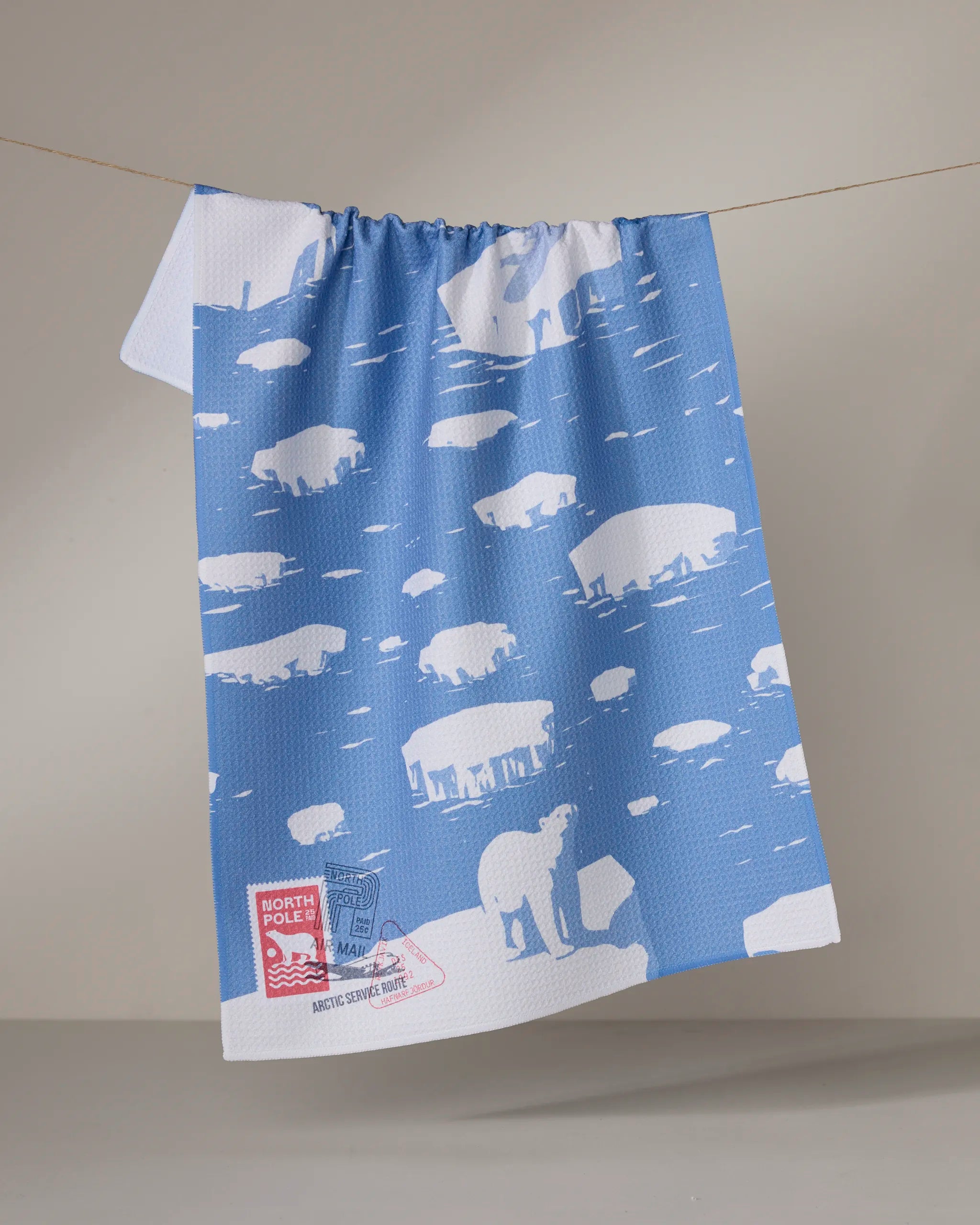 The Geometry Polar Float is a blue and white tea towel with polar bears on ice floes, hanging on a clothesline. Ultra-absorbent and made from recycled materials, it features a North Pole label with a polar bear graphic on the corner.