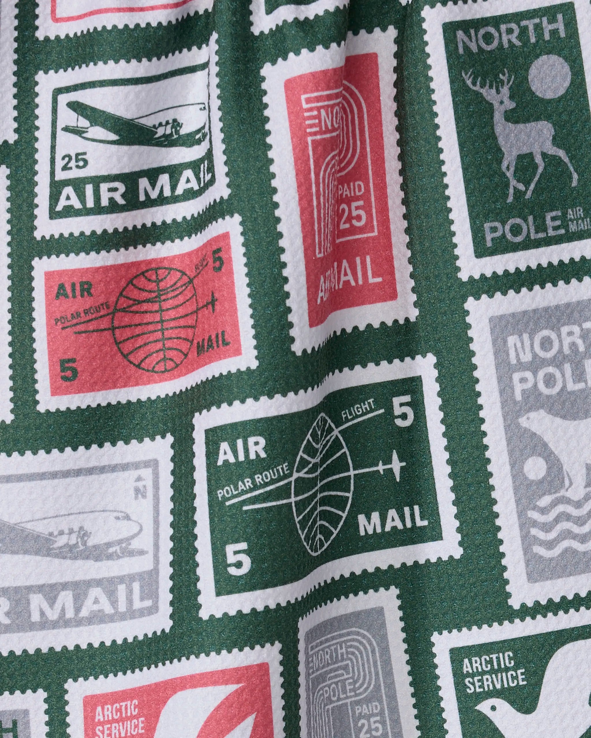 A close-up of Geometry’s Polar Post tea towel, patterned with vintage postage stamps—airplanes, a North Pole deer, AIR MAIL and POLAR ROUTE text—in green, red, and white. Quick-drying and made from recycled materials.