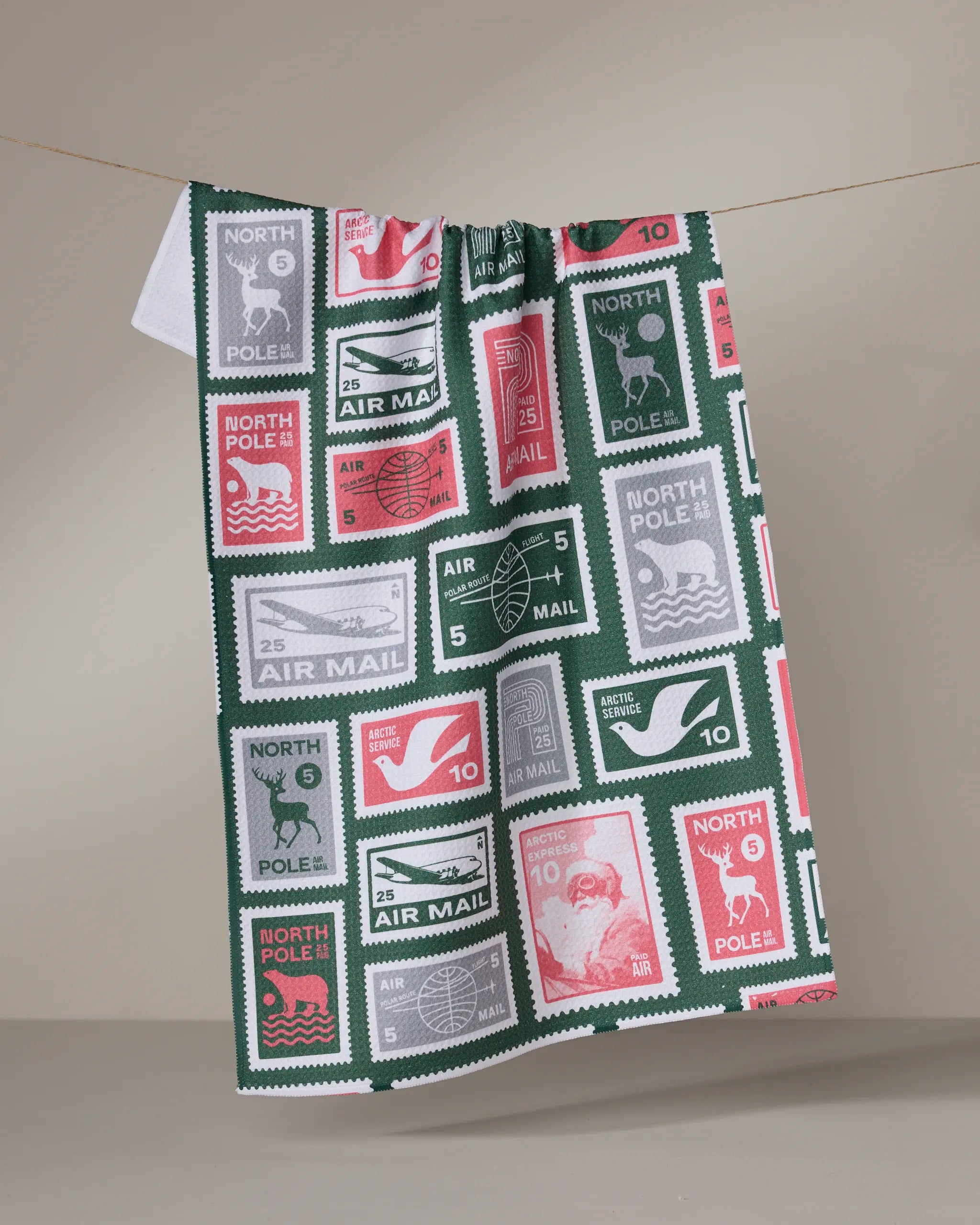 Geometry’s Polar Post tea towel features a quick-drying recycled fabric with vintage-style holiday and North Pole postage stamp designs in red, green, white, and gray, shown hanging on a line against a neutral background.
