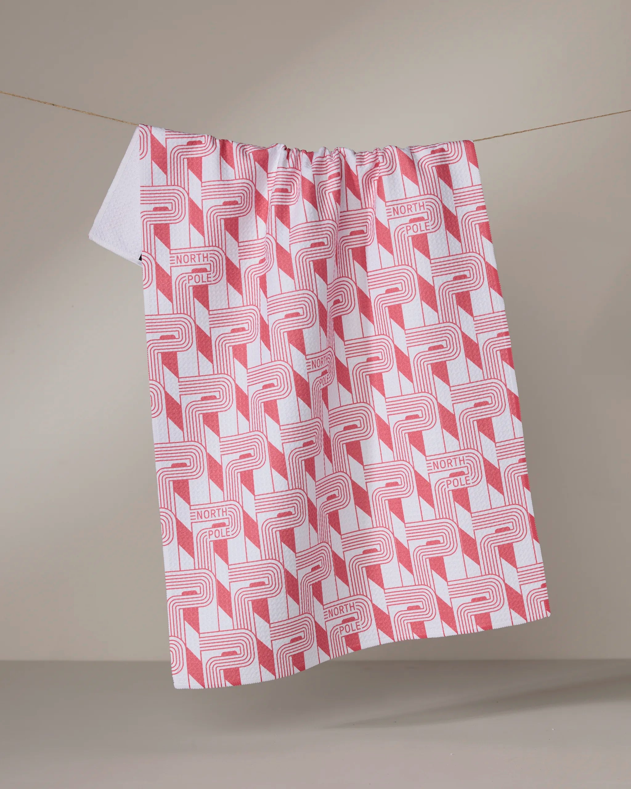 The Geometry Polemark towel, made from recycled materials, showcases a red geometric pattern and repeating "NORTH POLE" text on white fabric, draped over a string with a plain beige backdrop.