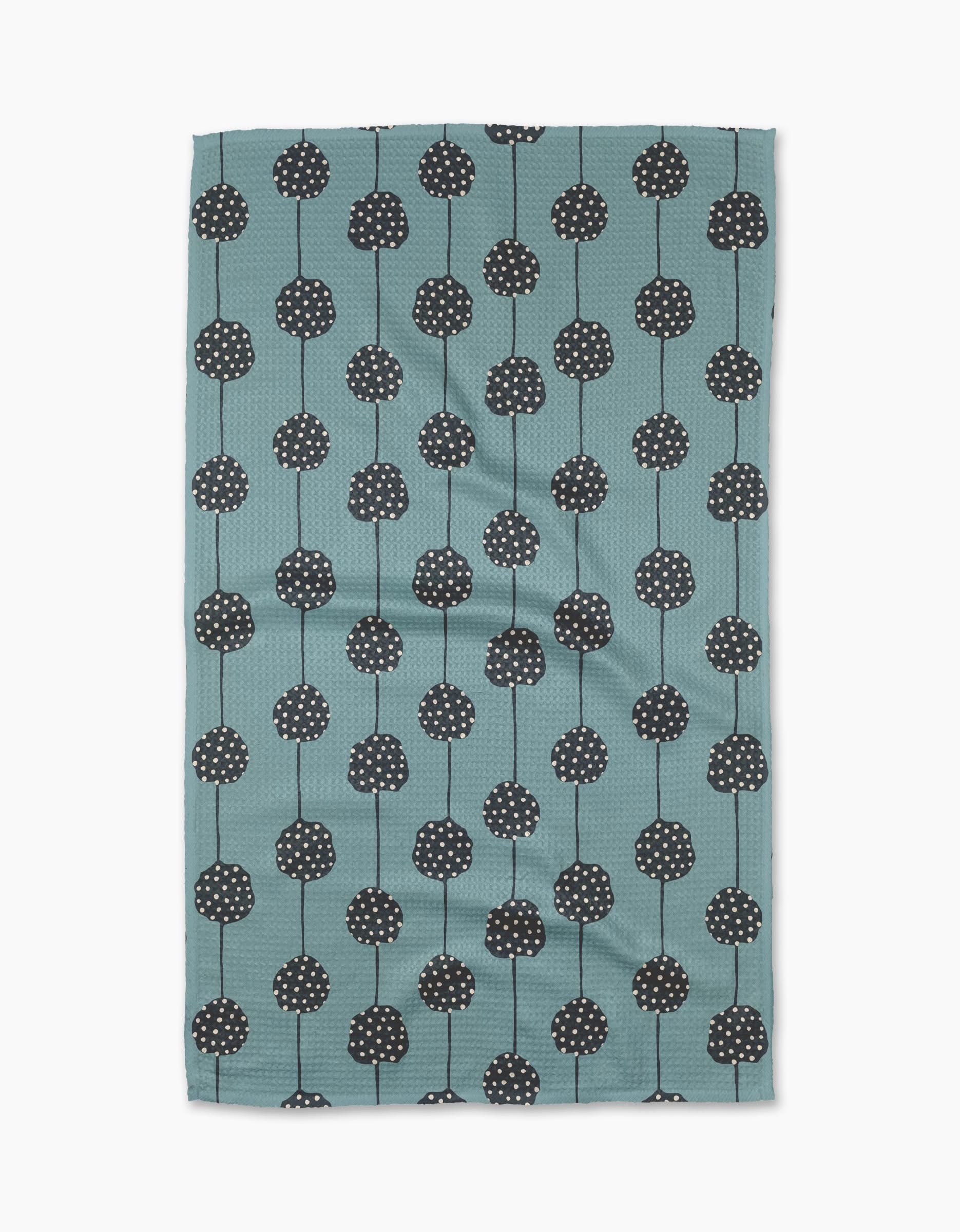 The Polka Dot Garland tea towel by Lizzie Clark is made from recycled materials in teal, with a pattern of black round flowers with white dots and thin stems arranged in vertical rows. The slightly wrinkled fabric is displayed on a white background.