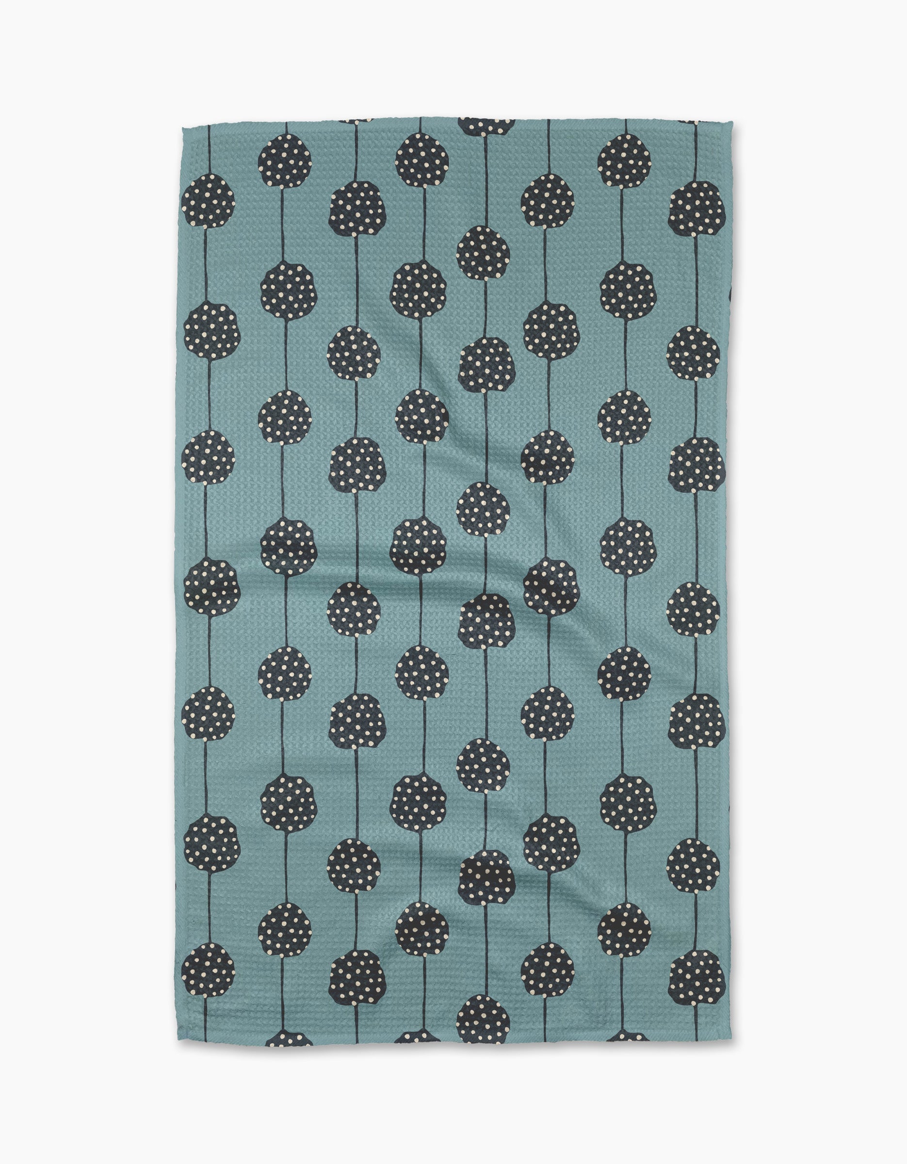The Polka Dot Garland tea towel by Lizzie Clark is made from recycled materials in teal, with a pattern of black round flowers with white dots and thin stems arranged in vertical rows. The slightly wrinkled fabric is displayed on a white background.