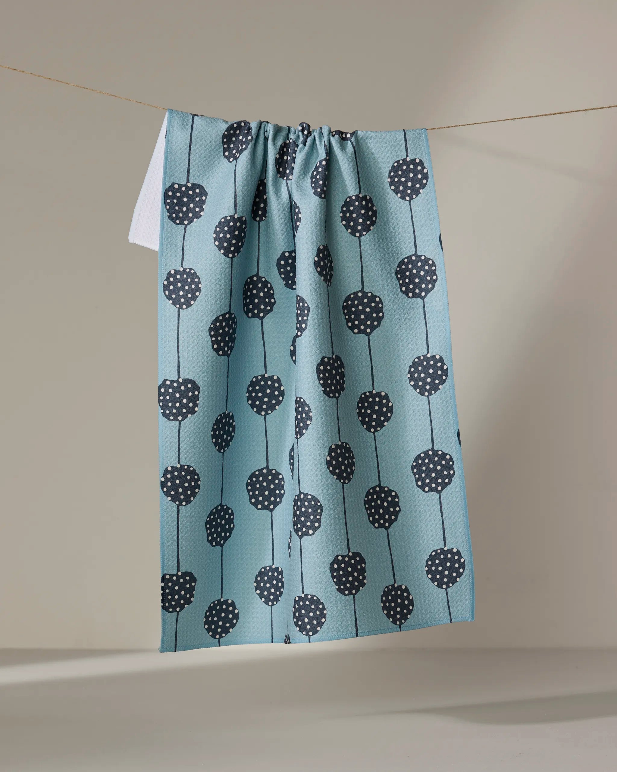 The Lizzie Clark Polka Dot Garland, an eco-friendly blue kitchen towel with dark blue dotted circles linked by vertical lines, hangs on a clothesline against a plain neutral background.