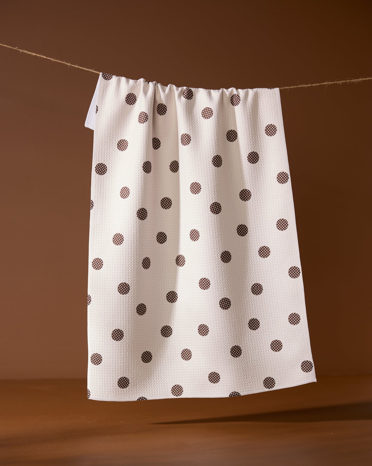 The Polka Picnic tea towel by Geometry, featuring black polka dots on white, hangs on a string against a brown background.