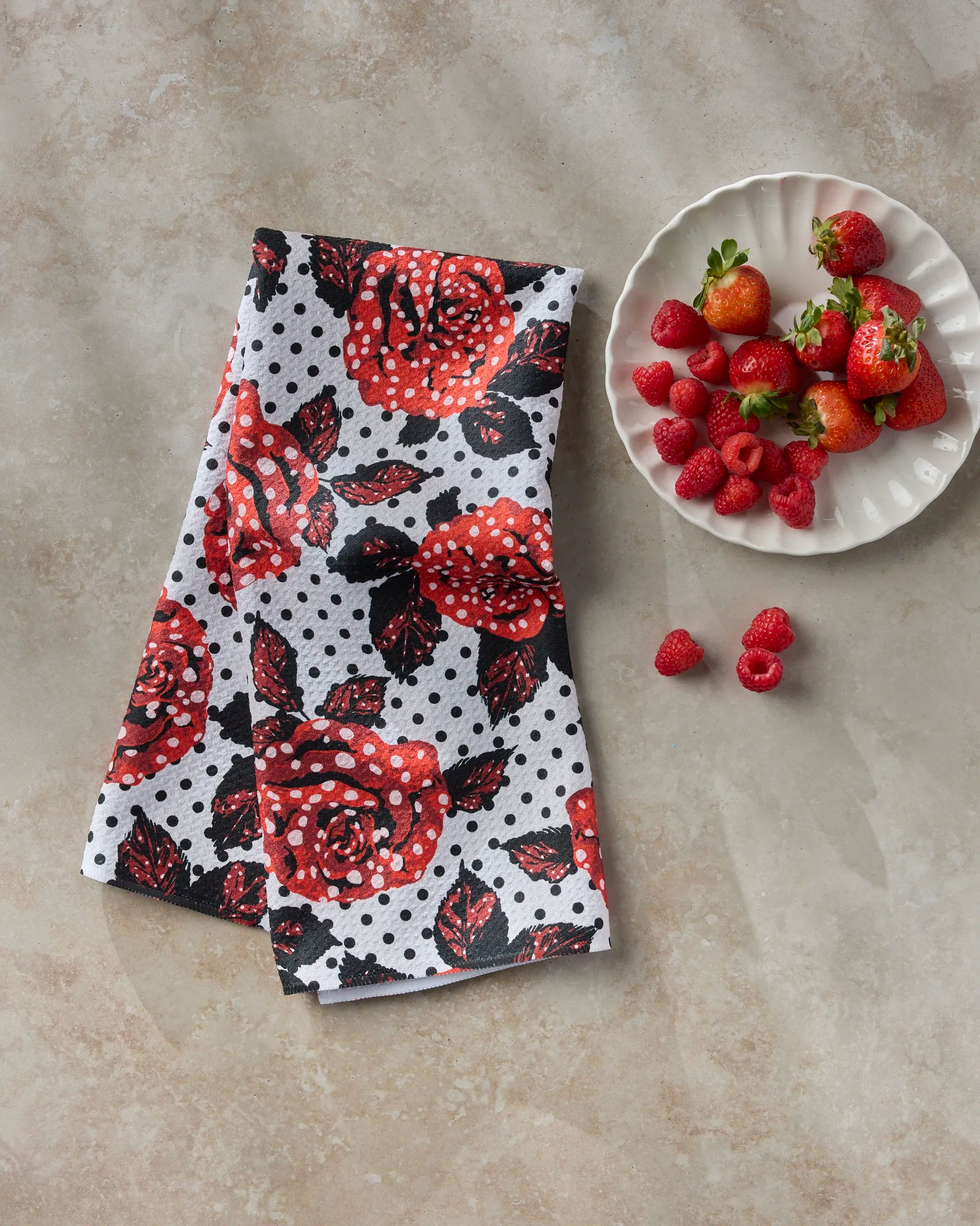 The Geometry Polka Rose recycled kitchen towel, featuring red roses and black polka dots on white, is placed on a light surface beside a white plate of fresh strawberries and raspberries.