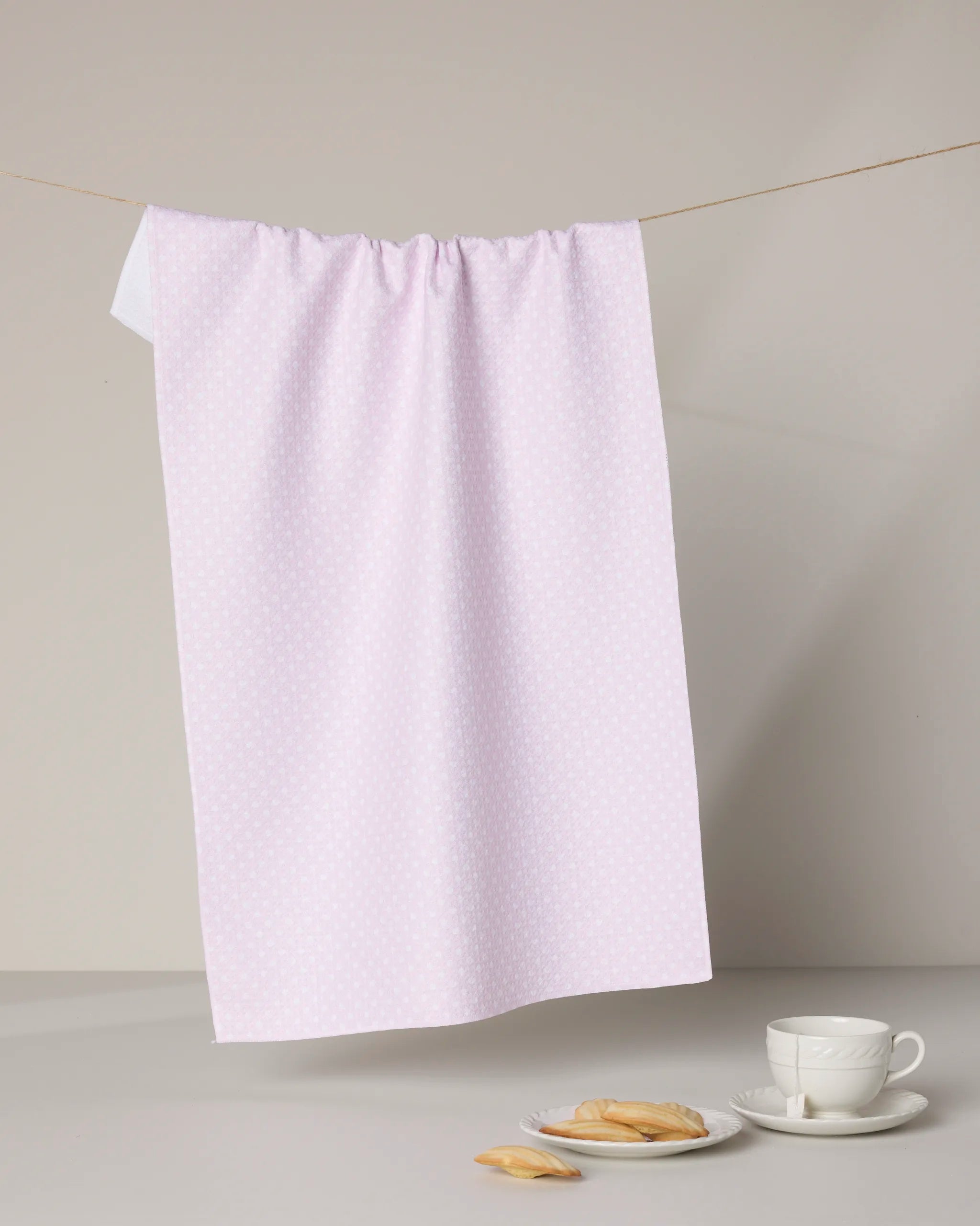 The Polka Val tea towel by Geometry, featuring a pale pink dotted design, hangs above a white teacup and cookies on a gray surface, showcasing the ultra-absorbent elegance of this stylish kitchen essential.