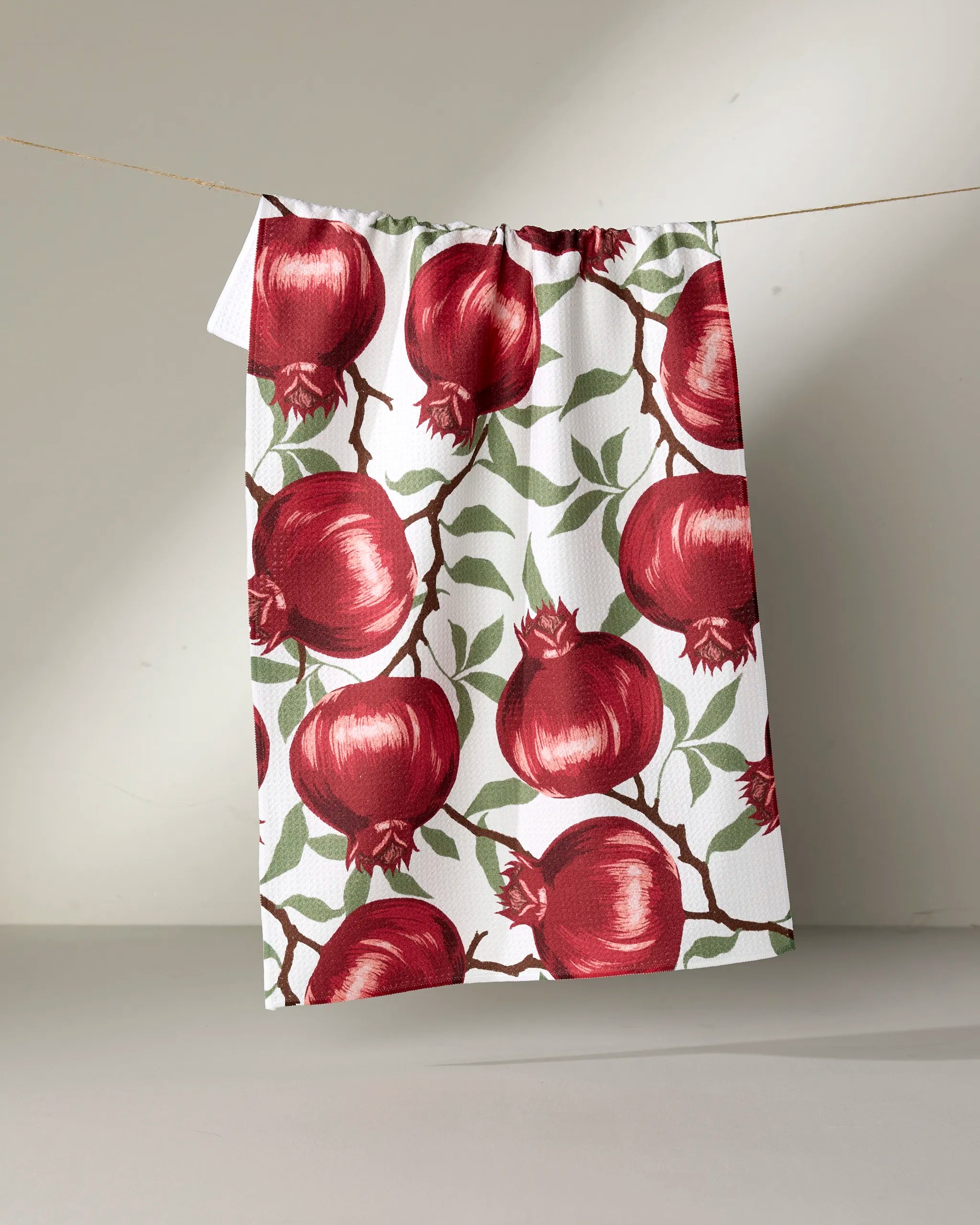 Ashley Reeves' Pomegranate Party tea towel, featuring bold red pomegranates and green leaves on white, hangs against a neutral background.