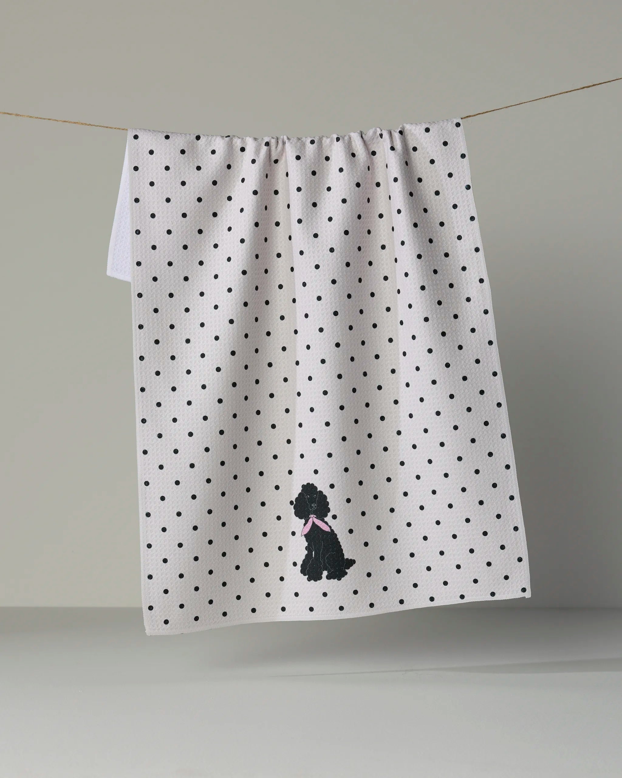 A white polka dot tea towel from Geometry’s Poodle Charm collection hangs on a line, showcasing an embroidered small black poodle with a pink bow near the bottom center. This ultra-absorbent towel is ideal for drying dishes while adding a charming touch to your kitchen décor.