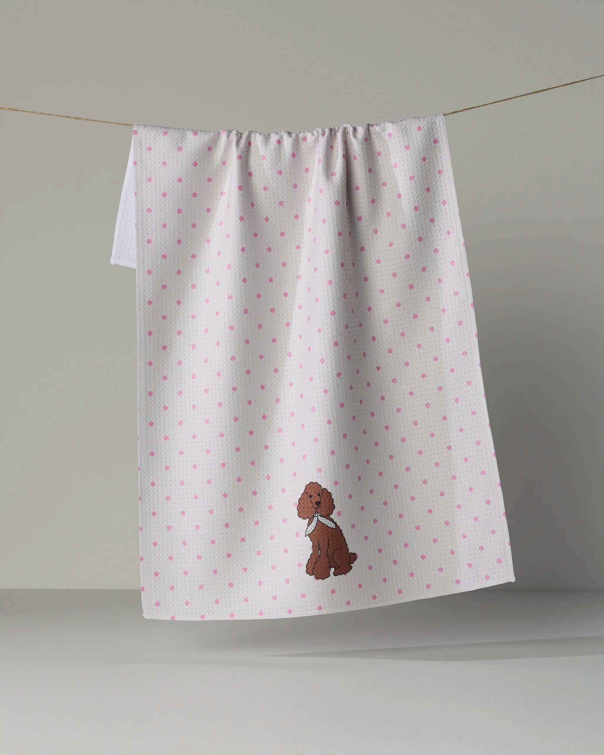 A white Geometry tea towel from the Poodle Charm collection, featuring pink polka dots and an embroidered brown poodle near the bottom, hangs on a line. The ultra-absorbent towel combines style and function.