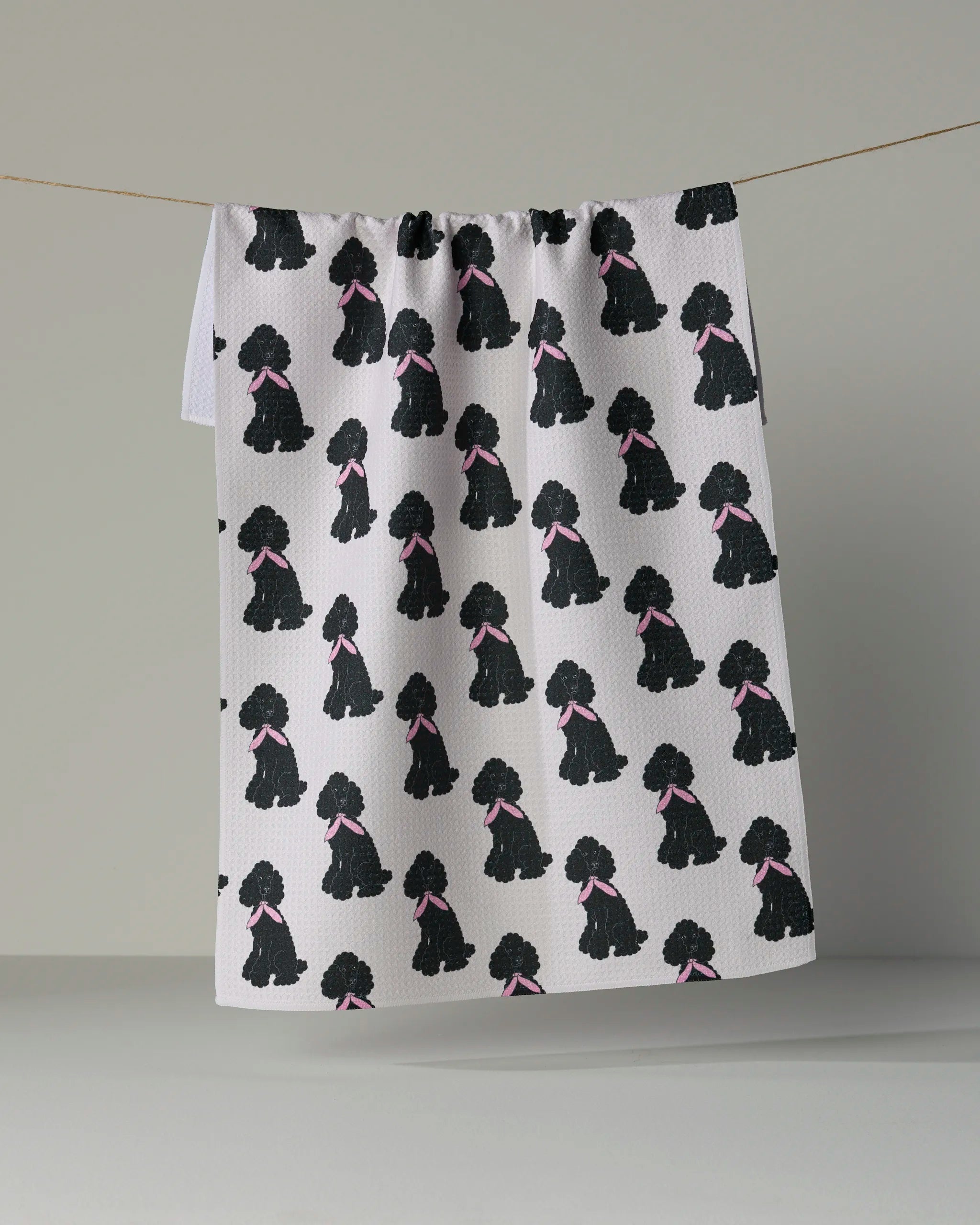 A white “Poodle Parade” tea towel by Geometry hangs on a line, featuring a repeating pattern of black poodles wearing pink bows.