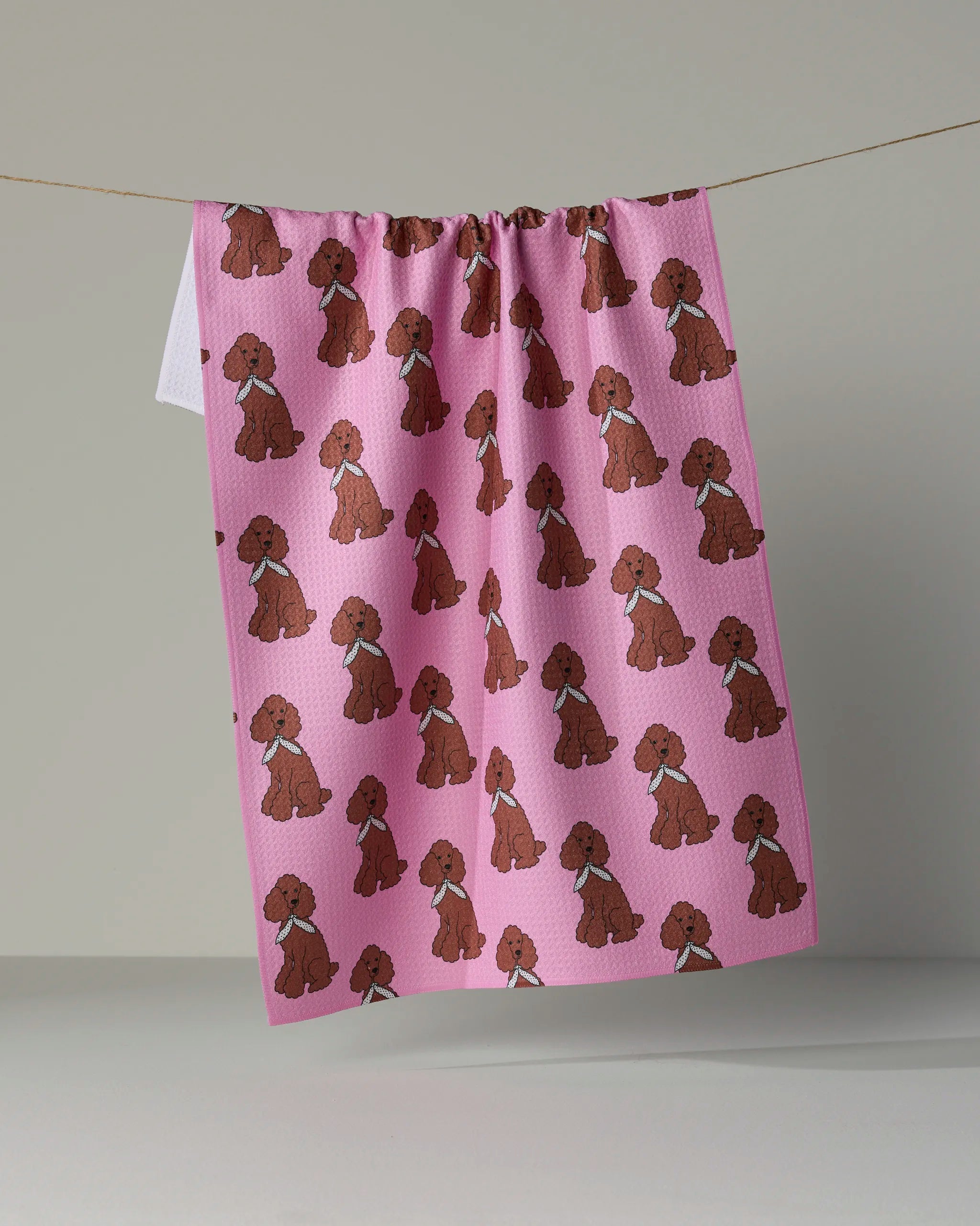 A “Poodle Parade” kitchen tea towel by Geometry hangs on a line, featuring a repeating pattern of brown cartoon poodles wearing blue collars against a pink background.