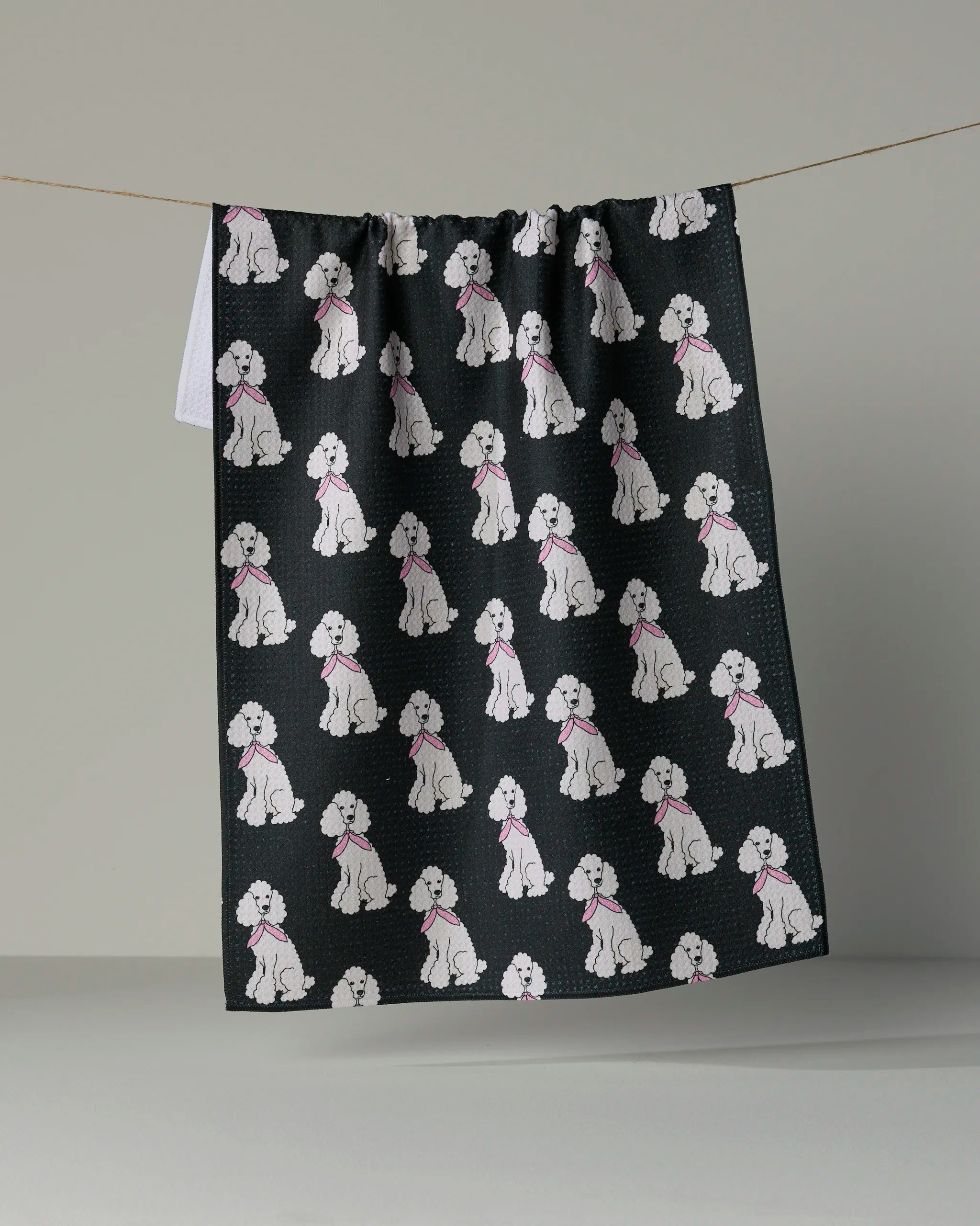 Introducing “Poodle Parade” by Geometry: a dark, absorbent tea towel adorned with a repeating pattern of white poodles in pink collars, shown hanging on a line against a neutral background.