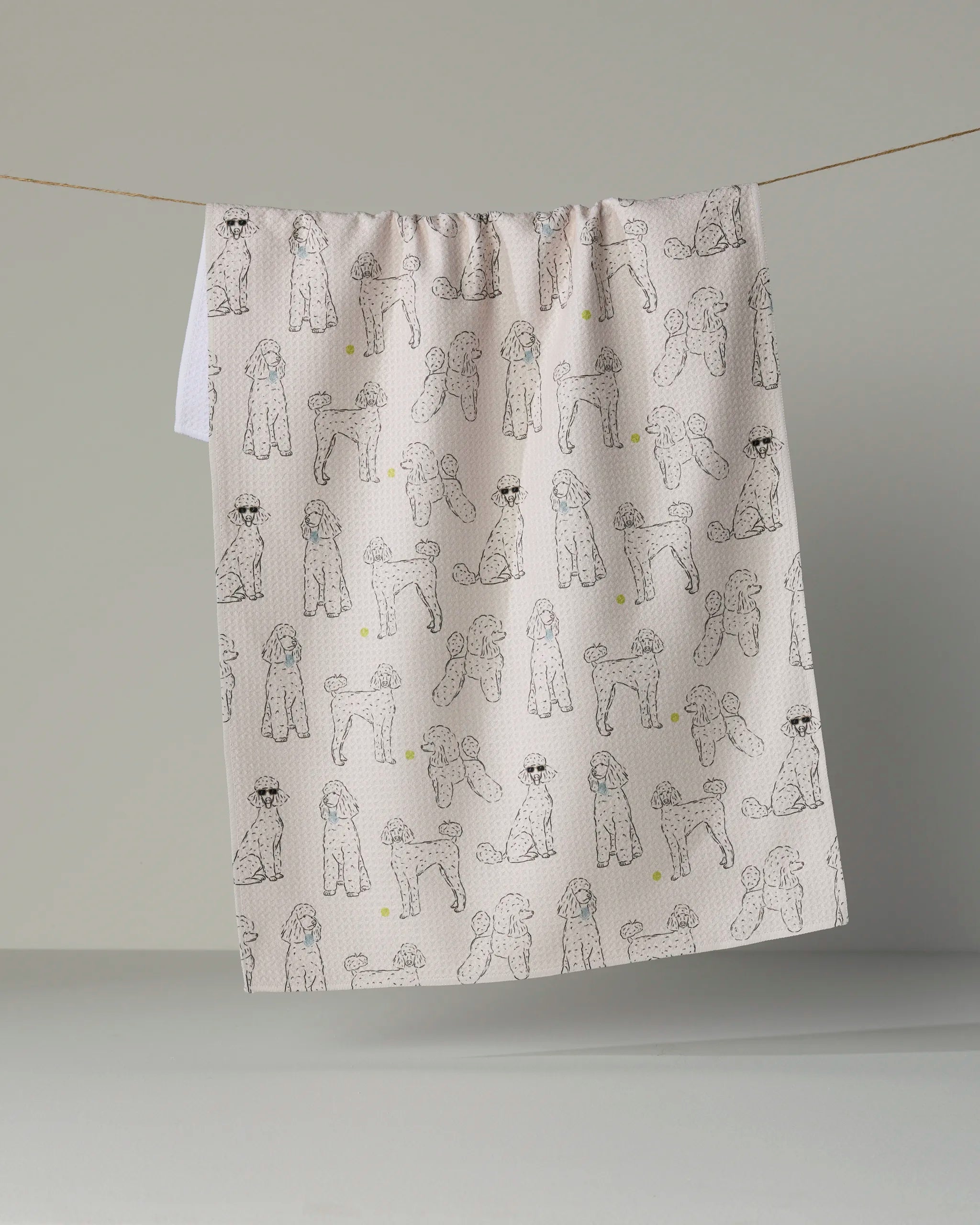 Geometry’s “Poodle Party” tea towel hangs on a line, showcasing a whimsical pattern of doodle-style poodles in various poses against a soft, dotted backdrop. This eco-friendly tea towel adds playful charm and style to your kitchen while providing the practical absorbency you need for everyday tasks.