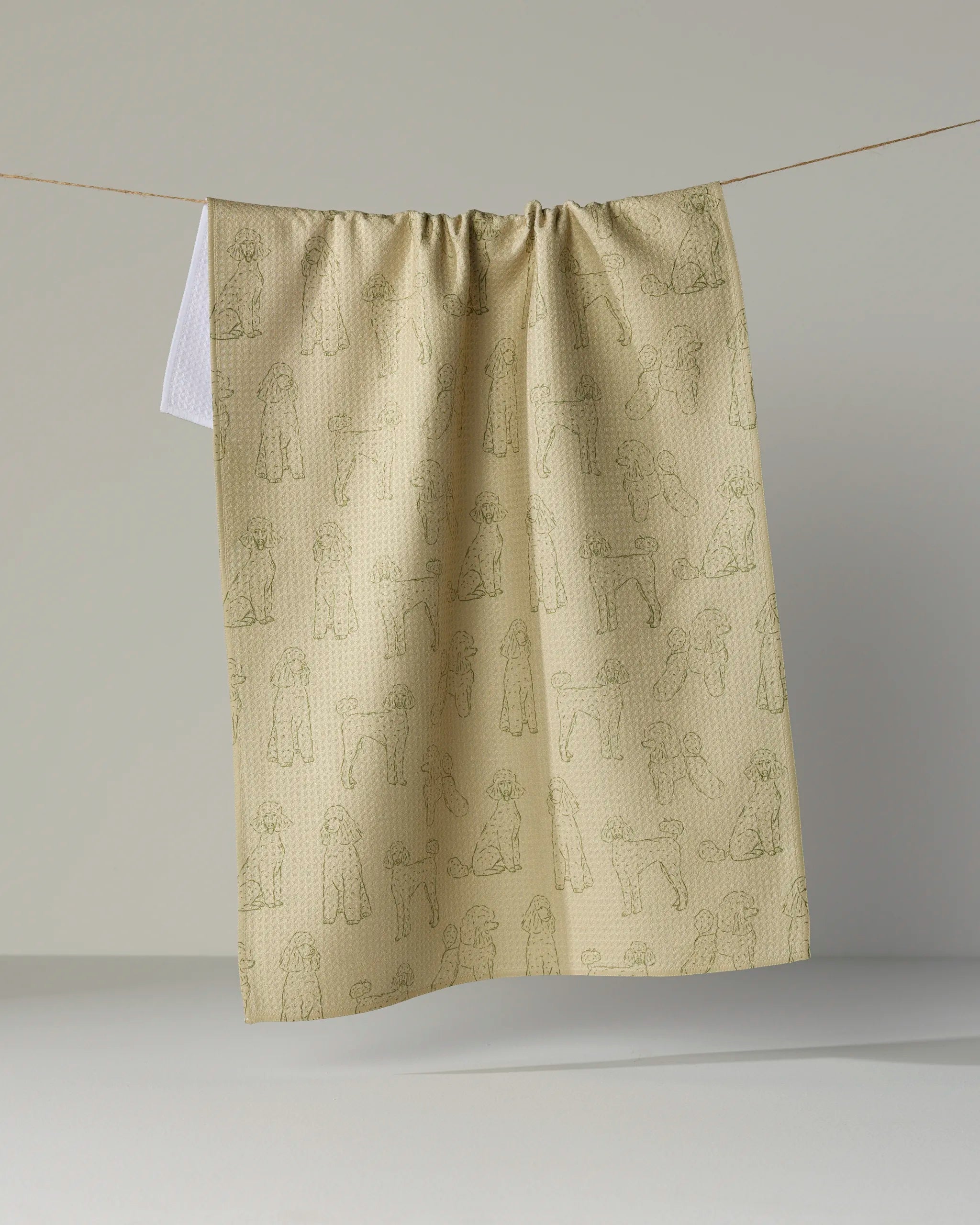 A beige, absorbent tea towel from Geometry called “Poodle Pups,” featuring a subtle line drawing pattern of people and dogs, hangs on a clothesline against a plain gray background.