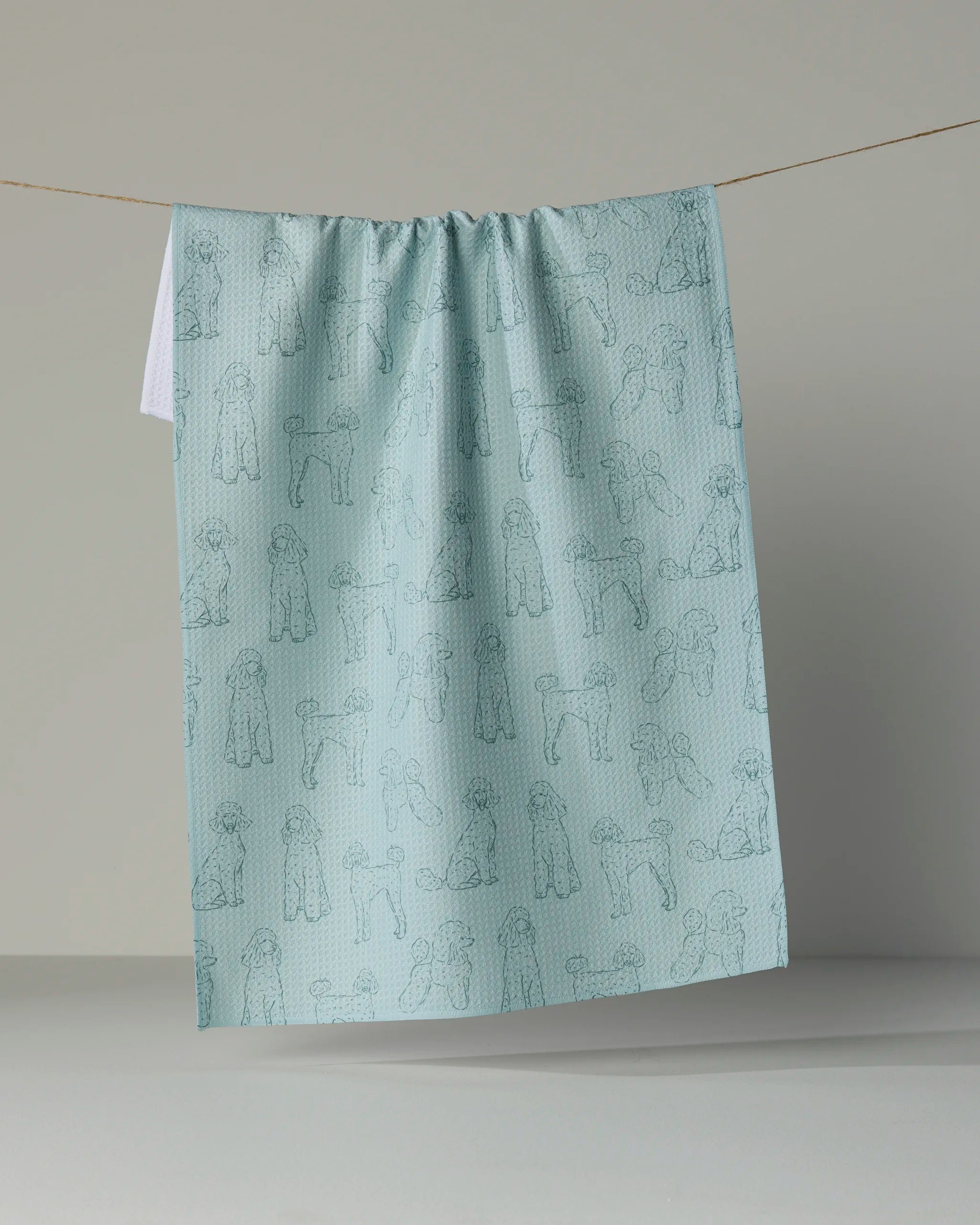 A light blue absorbent dish towel from Geometry, featuring line drawings of poodles from the Poodle Pups collection, hangs on a string against a plain, neutral background.