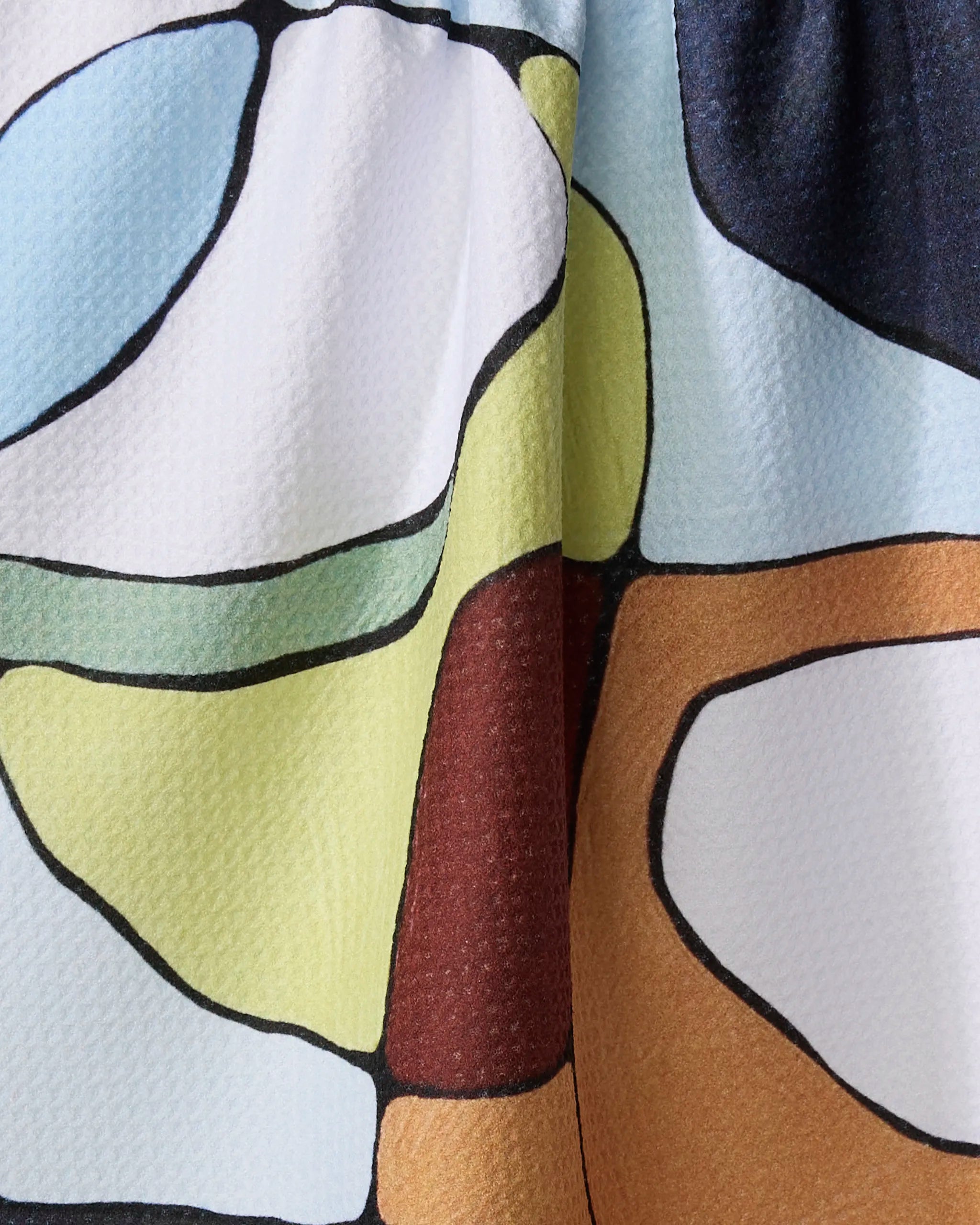 A close-up of the Rachel Austin "Poppies on Blue" kitchen tea towel, made from recycled materials and featuring a colorful abstract pattern with outlines and shades of yellow, blue, white, brown, and green on a textured surface.