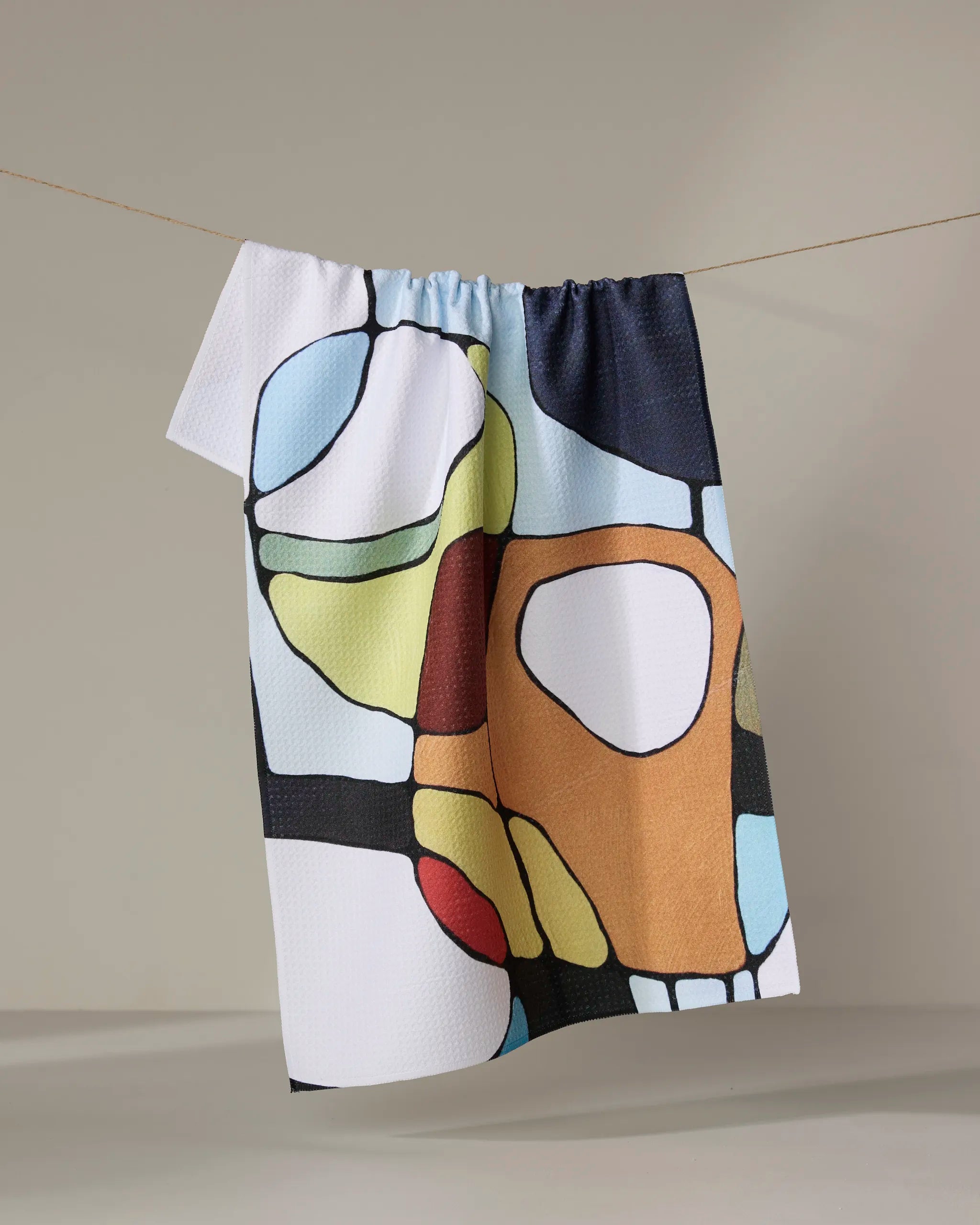 The "Poppies on Blue" kitchen tea towel by Rachel Austin features an abstract, stained glass-style poppy pattern and hangs on a line against a plain background—crafted from recycled materials.