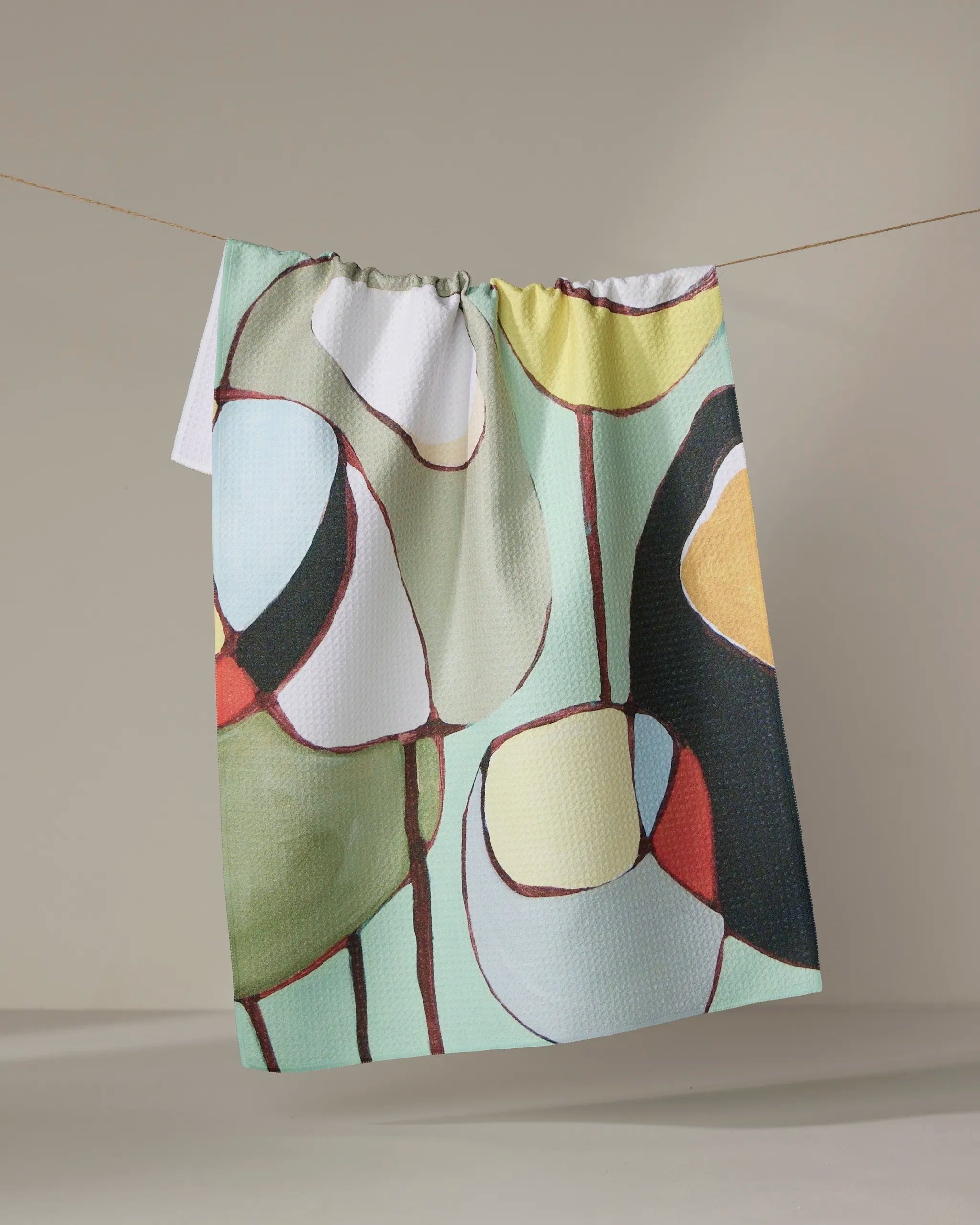 The "Poppies on Mint" tea towel by Rachel Austin, made from recycled materials, displays vibrant green, red, yellow, black, and white abstract shapes and hangs against a neutral background.