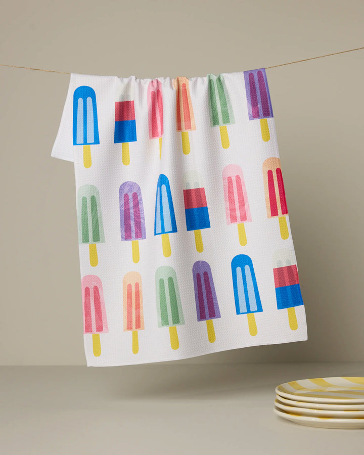 The Poppy Popsicles tea towel by Geometry features playful popsicle illustrations on white and hangs above a stack of yellow and white striped plates—perfect for adding a cheerful touch to your kitchen.