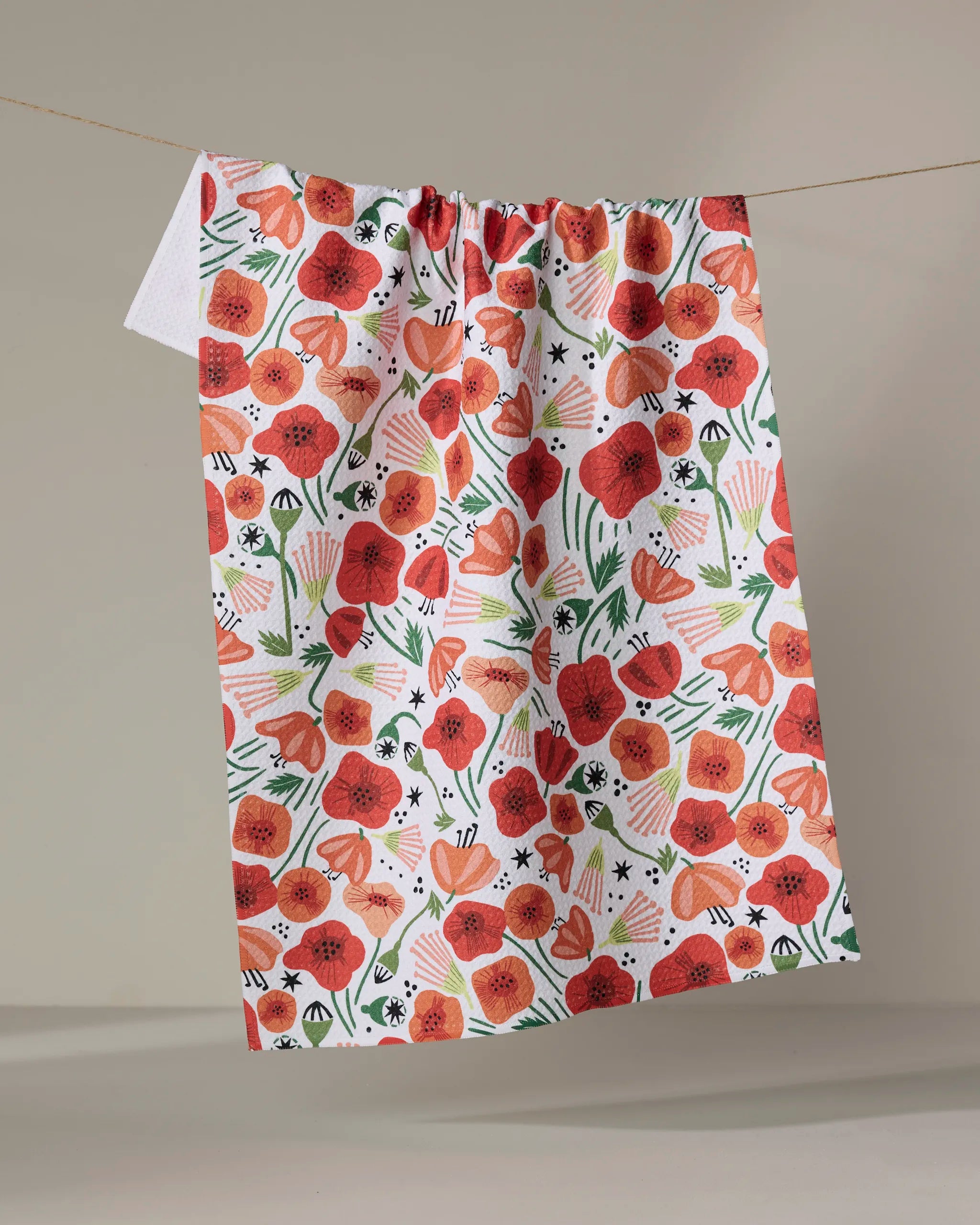 The "Poppy Power" quick-drying towel by Elisabeth Wing features vibrant red and orange poppies, green leaves, and abstract shapes, displayed on a clothesline against a neutral background.